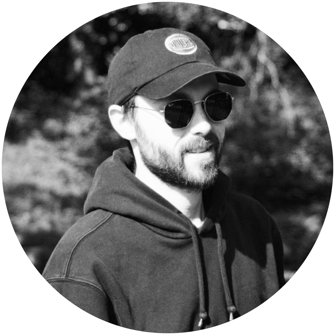 A man with a beard wearing a baseball cap, sunglasses, and a hoodie outdoors.