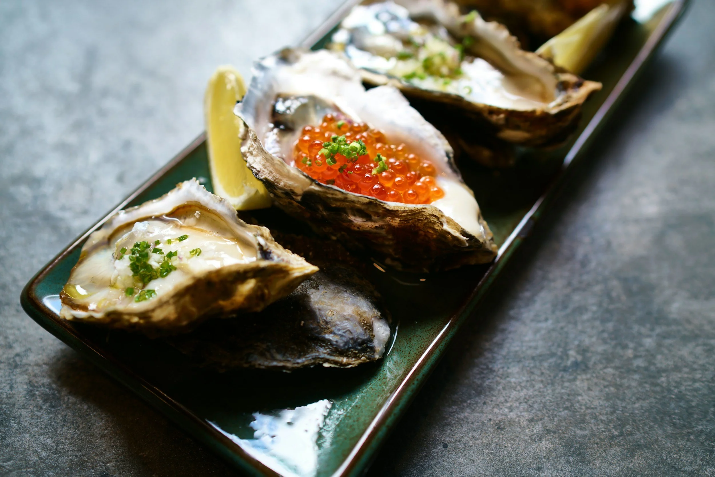 Close-up of oysters on a platter, topped with various garnishes such as salmon roe, chopped herbs, and lemon wedges, served on a dark plate.
