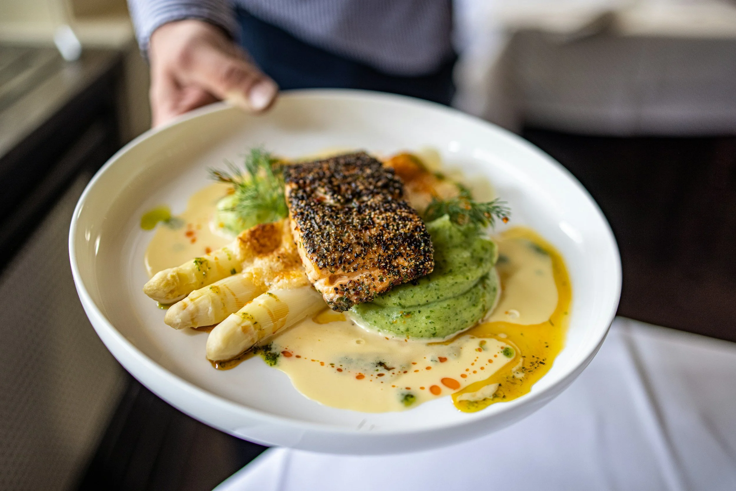 A plated dish featuring a piece of seared fish crusted with black sesame seeds, served with white asparagus, pureed green vegetables, and drizzled with a yellow sauce, garnished with fresh herbs.