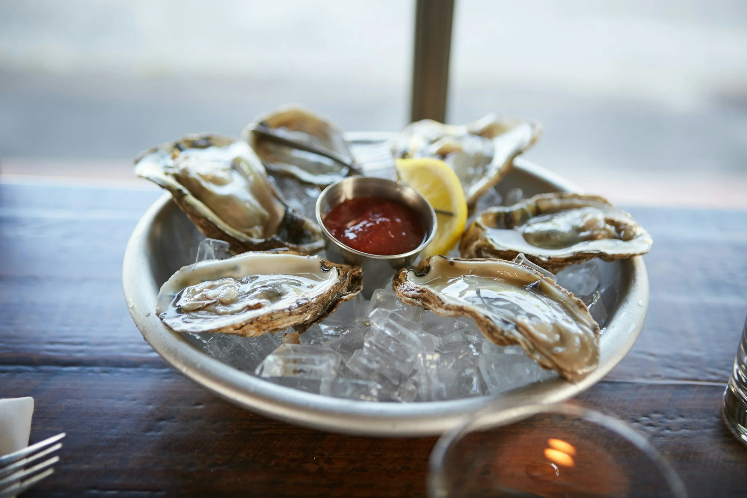 A white bowl of raw oysters on ice with lemon wedges and a cup of cocktail sauce, set on a wooden table.