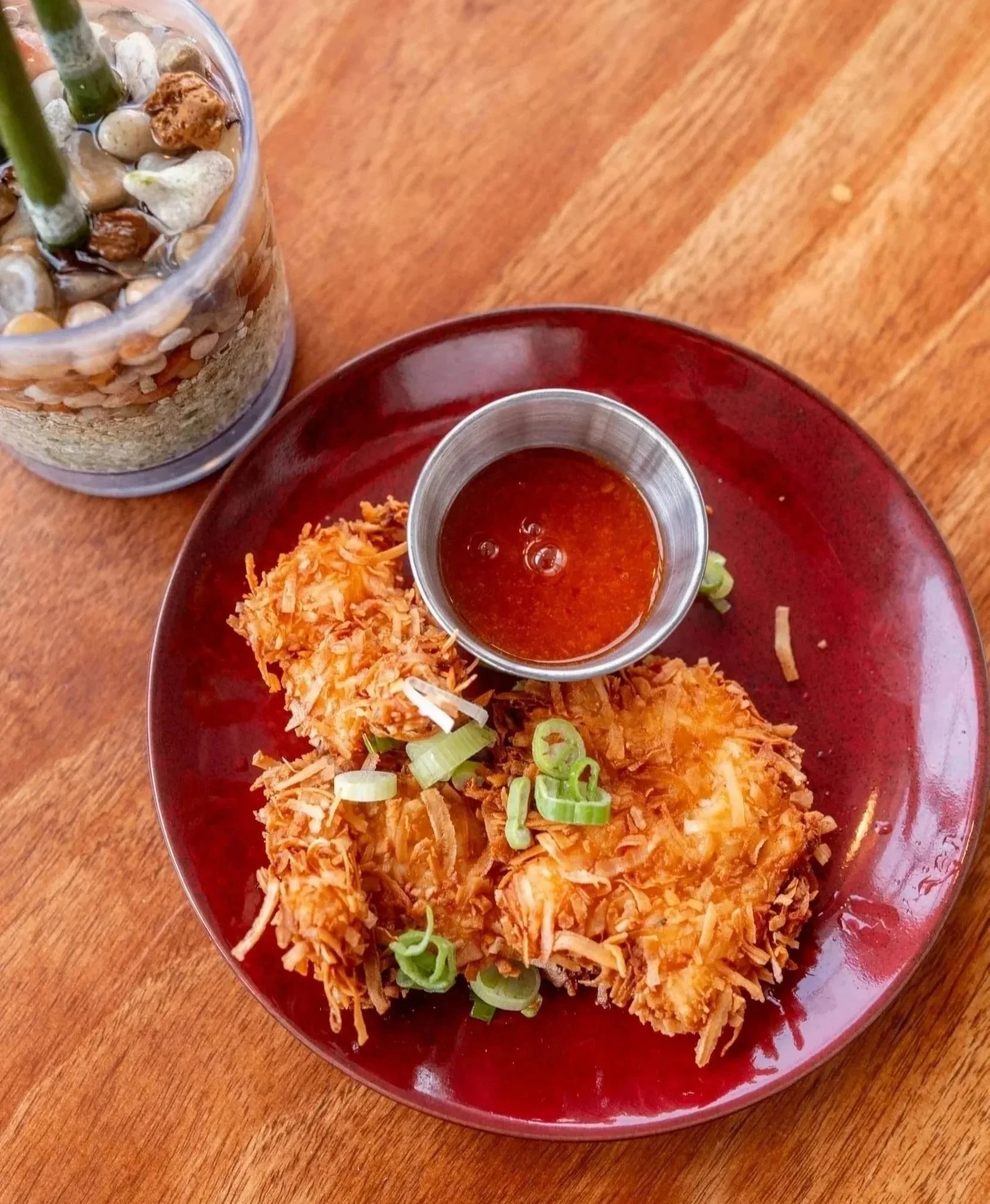A red plate with seasoned fried shredded chicken topped with chopped green onions, served with a small cup of red dipping sauce on a wooden table, and a glass jar containing pebbles and plant stems in the background.