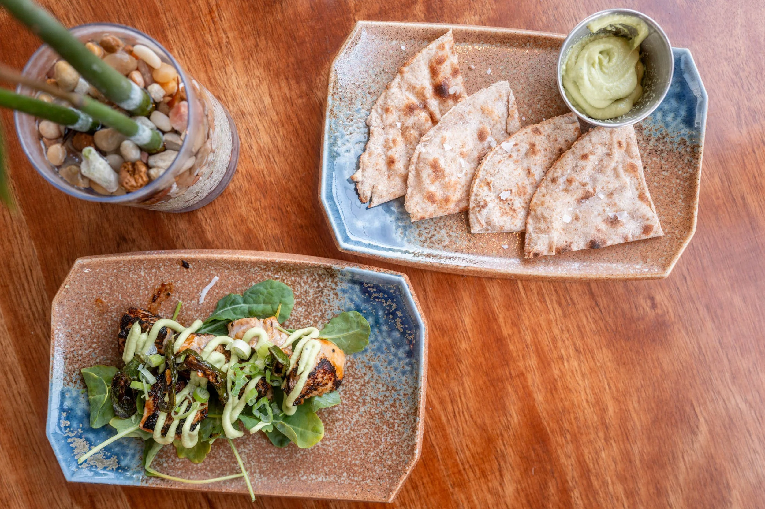 A plate of quesadillas with a side of guacamole, a bowl of salad, and a glass with green straws on a wooden table.