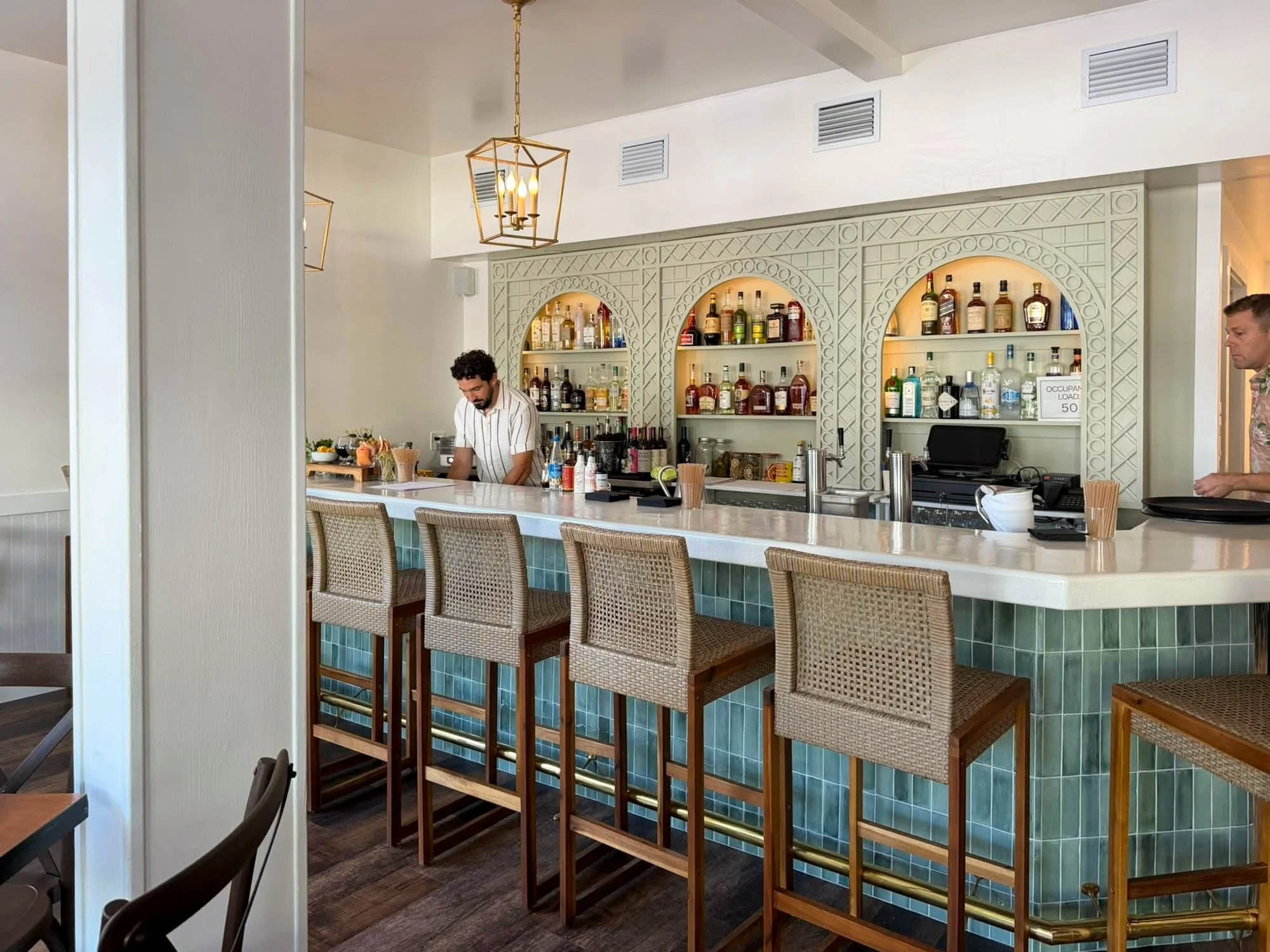 Indoor bar with a white countertop and wicker barstools, decorative shelving with assorted bottles of alcohol, two pendant lights hanging from the ceiling, a bartender preparing drinks, and a customer on the right side of the image.