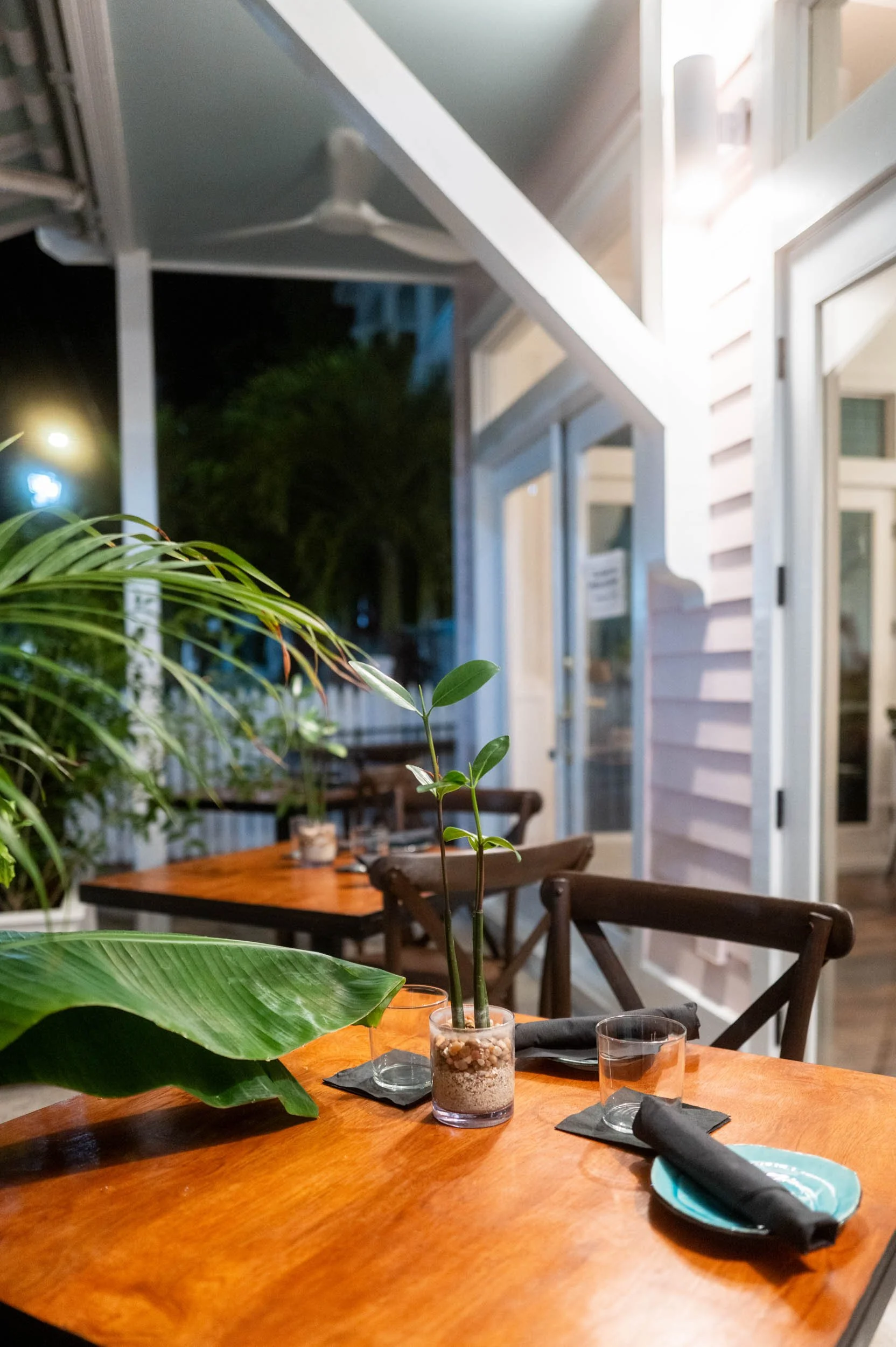 A wooden outdoor dining table set with black cloth napkins, glassware, and a decorative plant in a glass container. The setting is near a house with white siding and large windows, with trees and outdoor lighting visible in the evening.