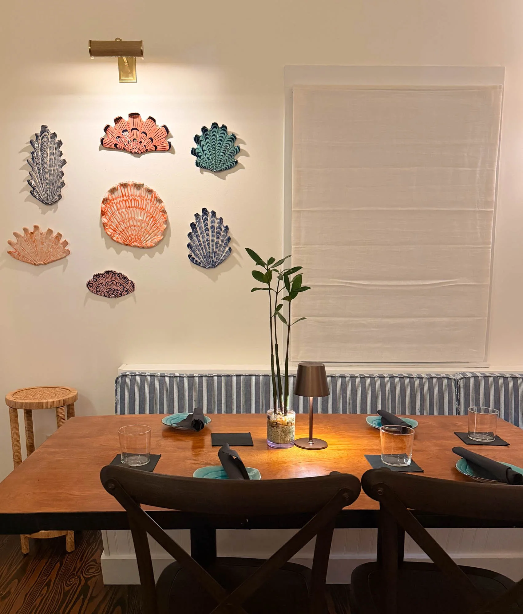 A dining room table set with four glasses, teal napkins, and teal plates. A centerpiece with a green plant in a glass container filled with stones. The table has a small table lamp and a placemat. The background has a wall with decorative seashells a