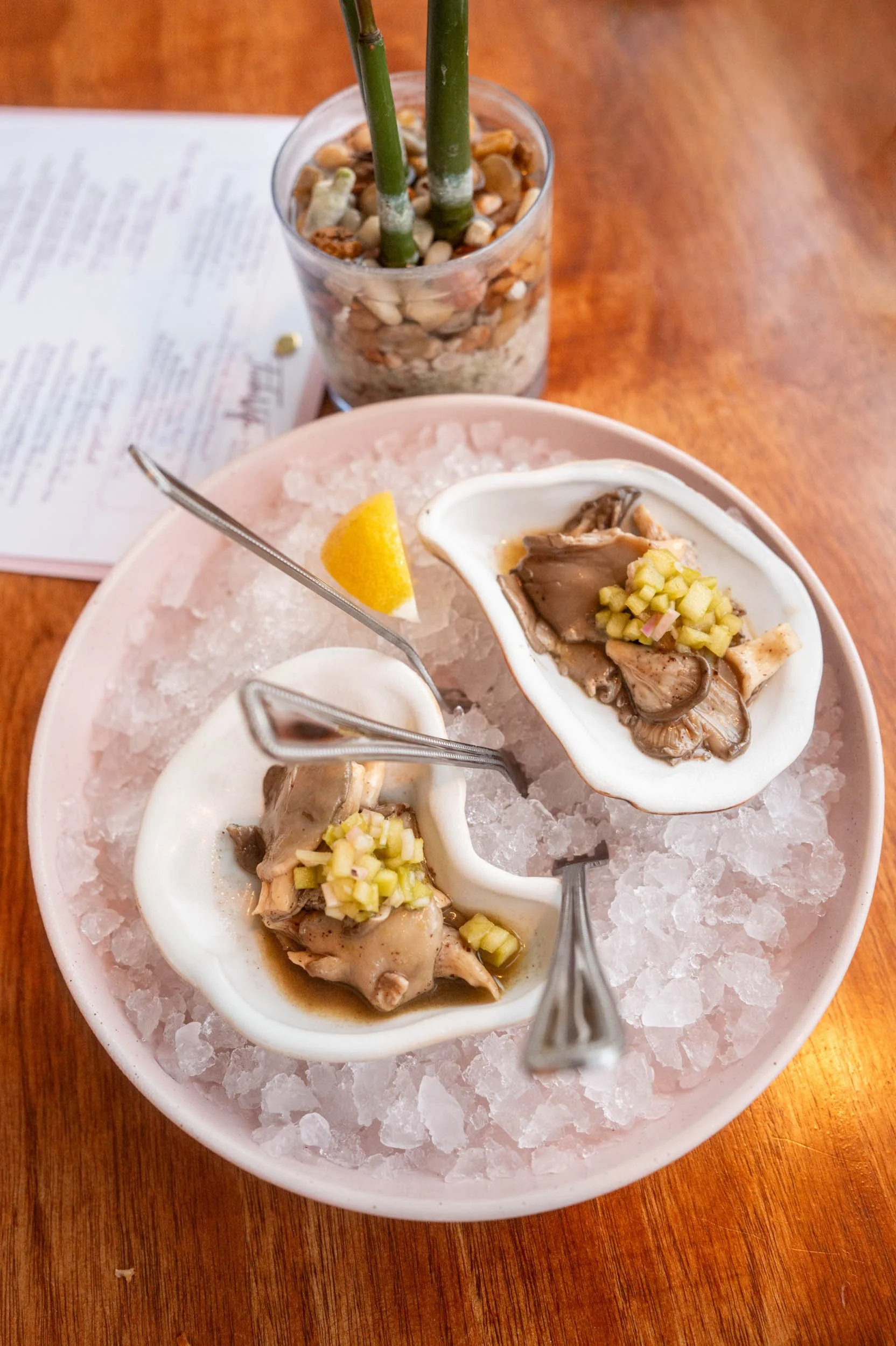Close-up of a white oval dish with oysters topped with chopped green onions, placed on a bed of crushed ice on a wooden table. In the background, a glass of water with rocks and bamboo stalks, and a menu or paper are visible.
