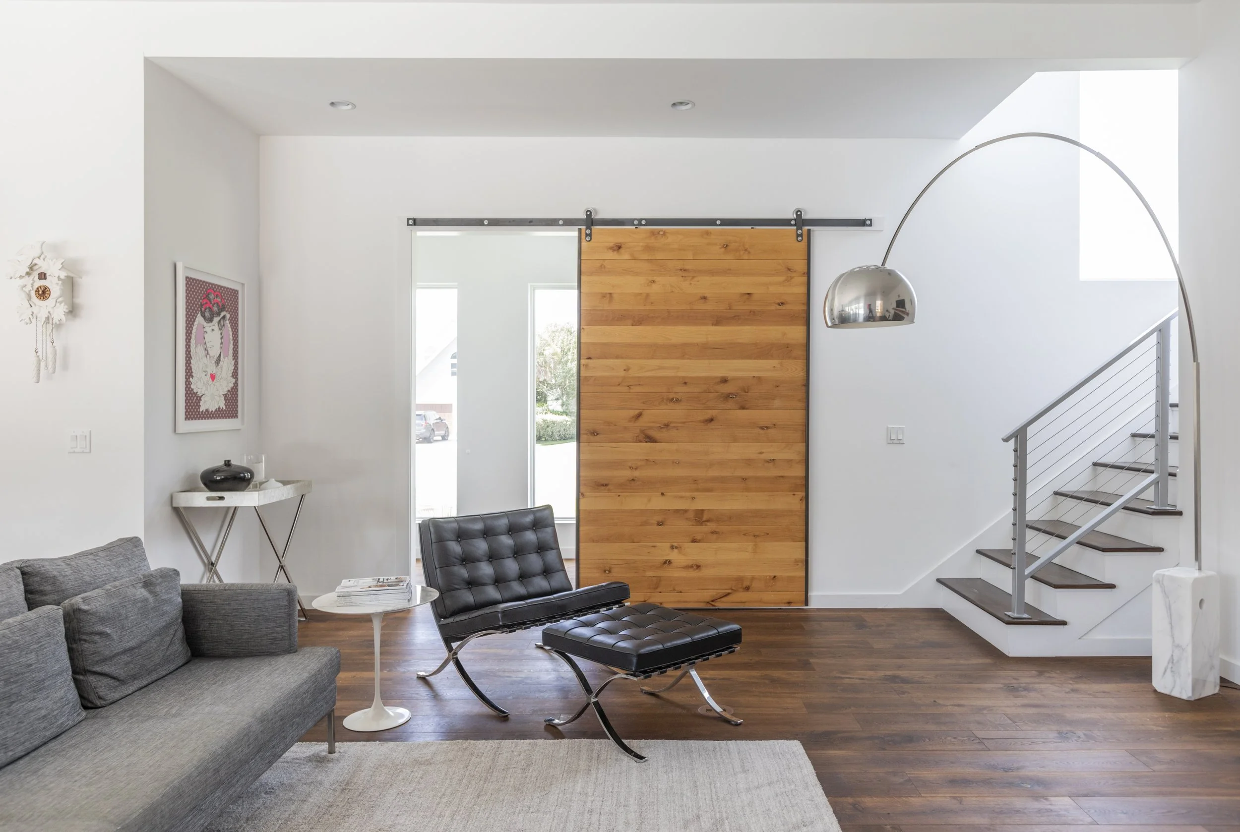 Modern living room with gray sofa, black leather lounge chair, white side table, wooden sliding barn door, staircase, and artwork on white walls.