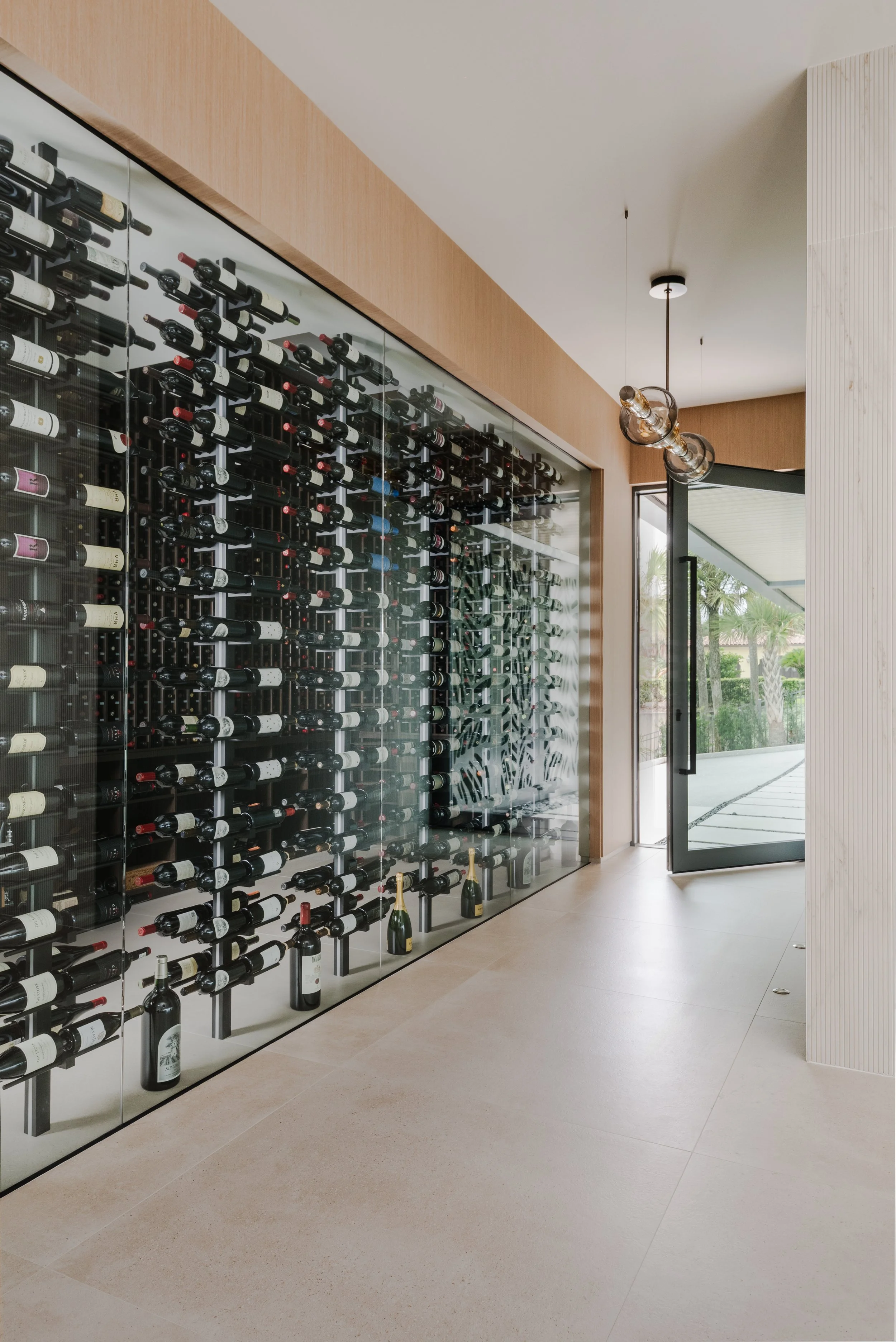 Modern wine cellar with a glass wall display of wine bottles and a large window showing greenery outside. The room features minimalist decor and pendant lighting.