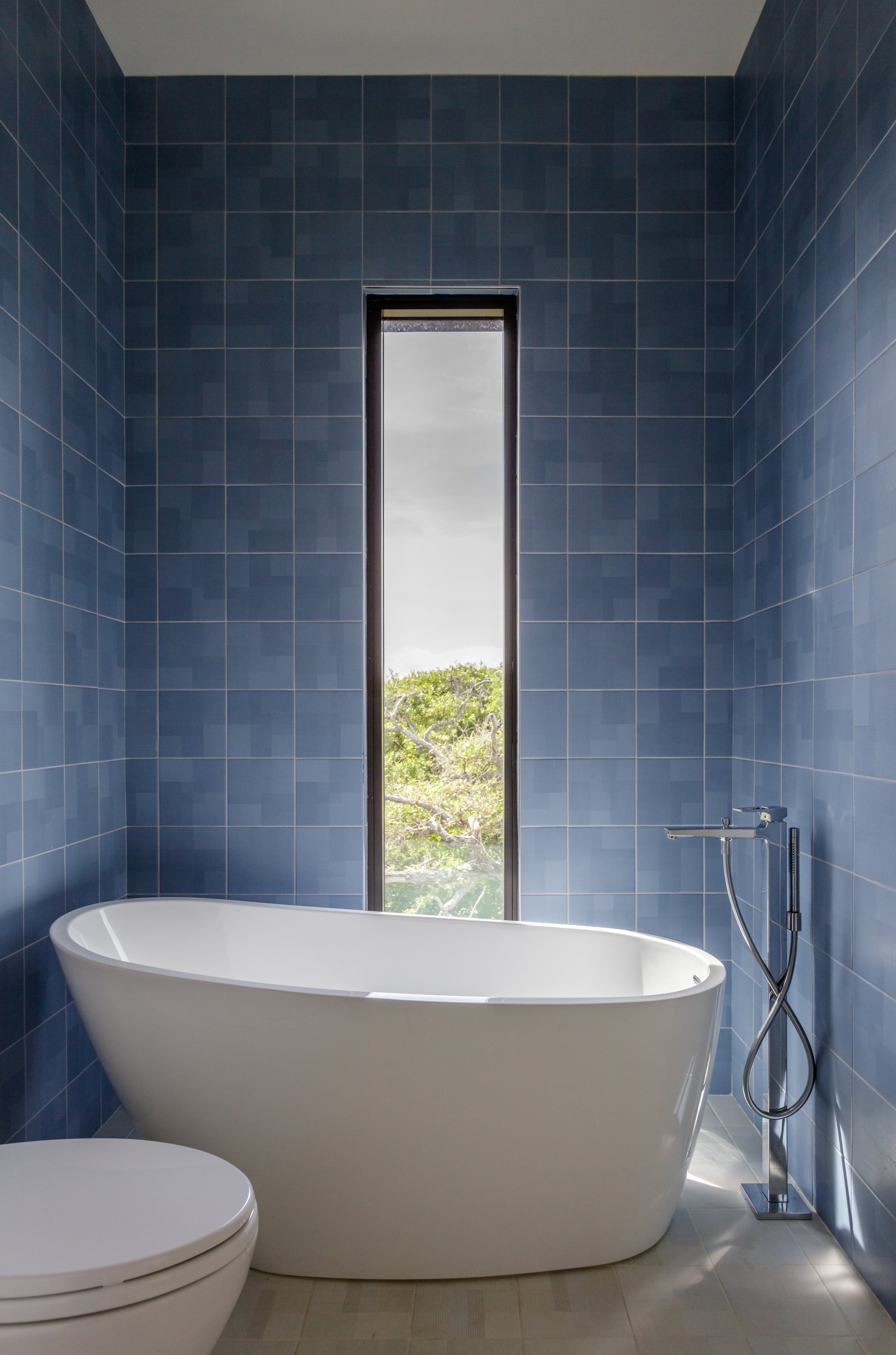 Modern bathroom with a white freestanding bathtub, blue tiled walls, a toilet, and a window showing green trees outside.