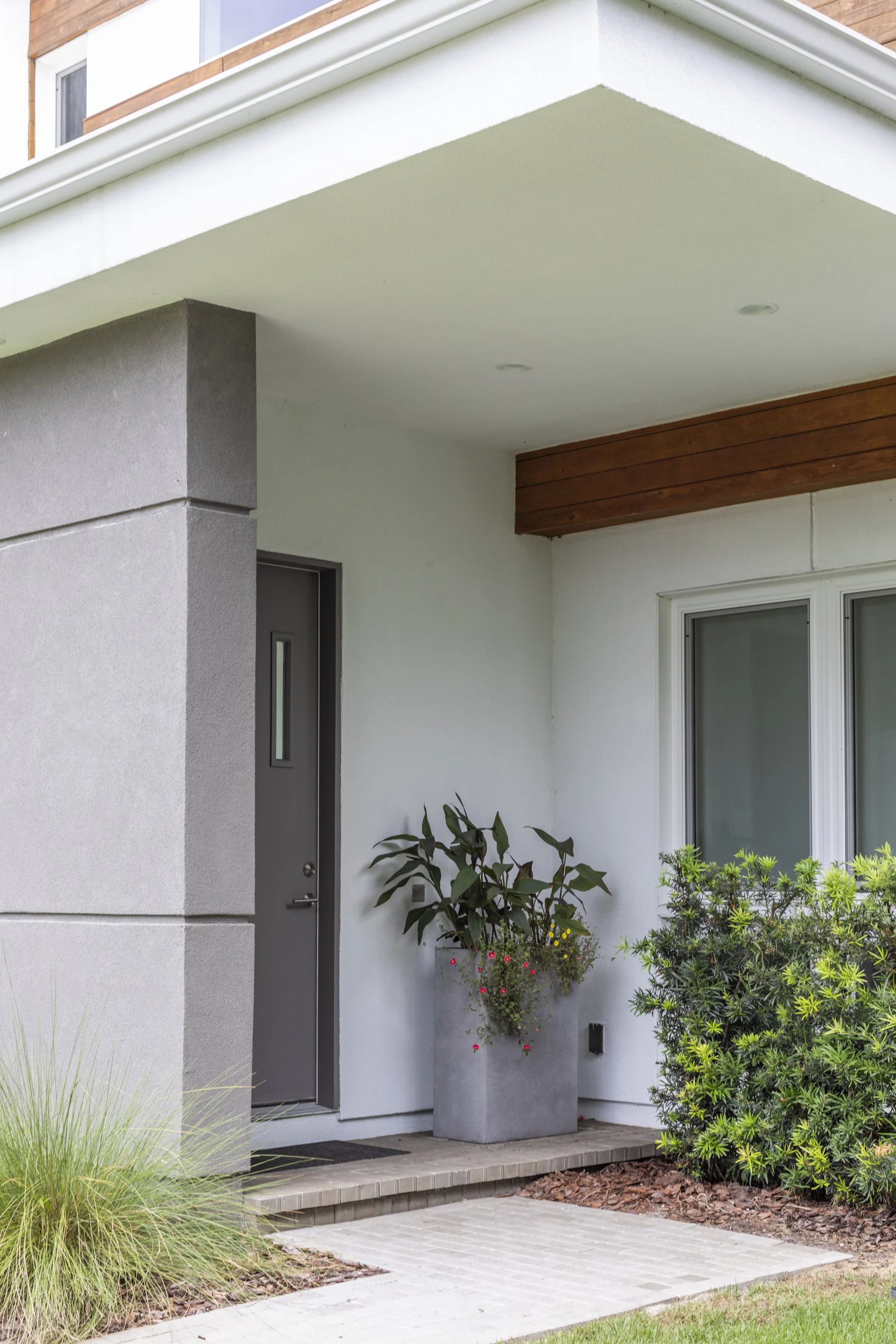 Modern house entrance with gray door, small window, white exterior walls, plants, and a brick pathway.