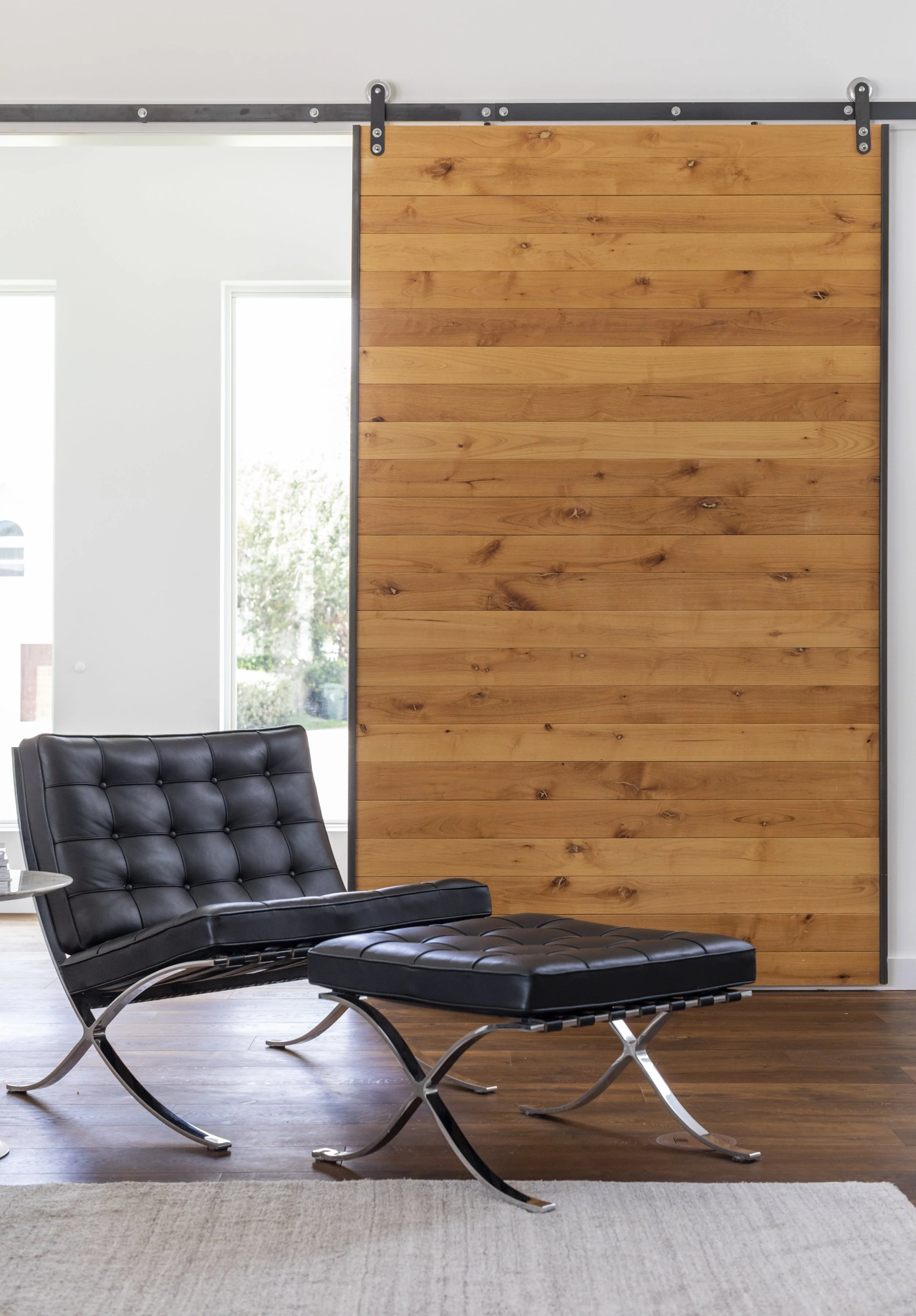 Modern black leather lounge chair and matching ottoman in front of a wooden sliding barn door, with a light-colored rug and hardwood floor in a bright room with large windows.