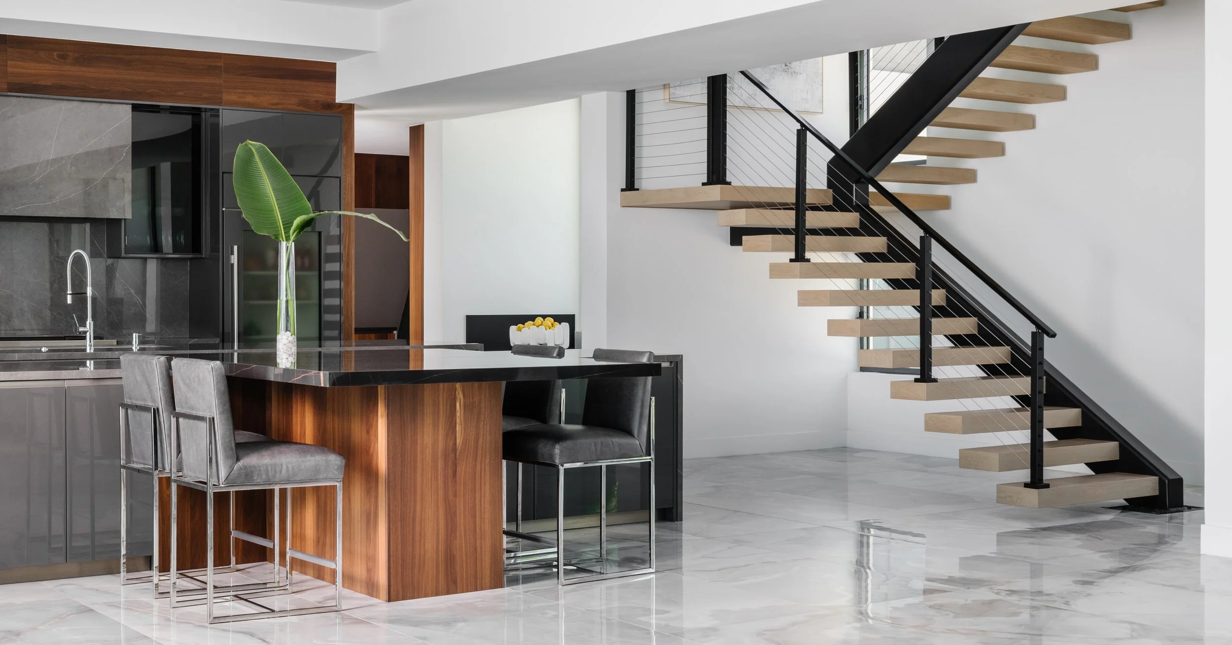 Modern interior with kitchen island, black countertops, gray barstools, a tall green plant, and a floating staircase with wooden steps and black railings.
