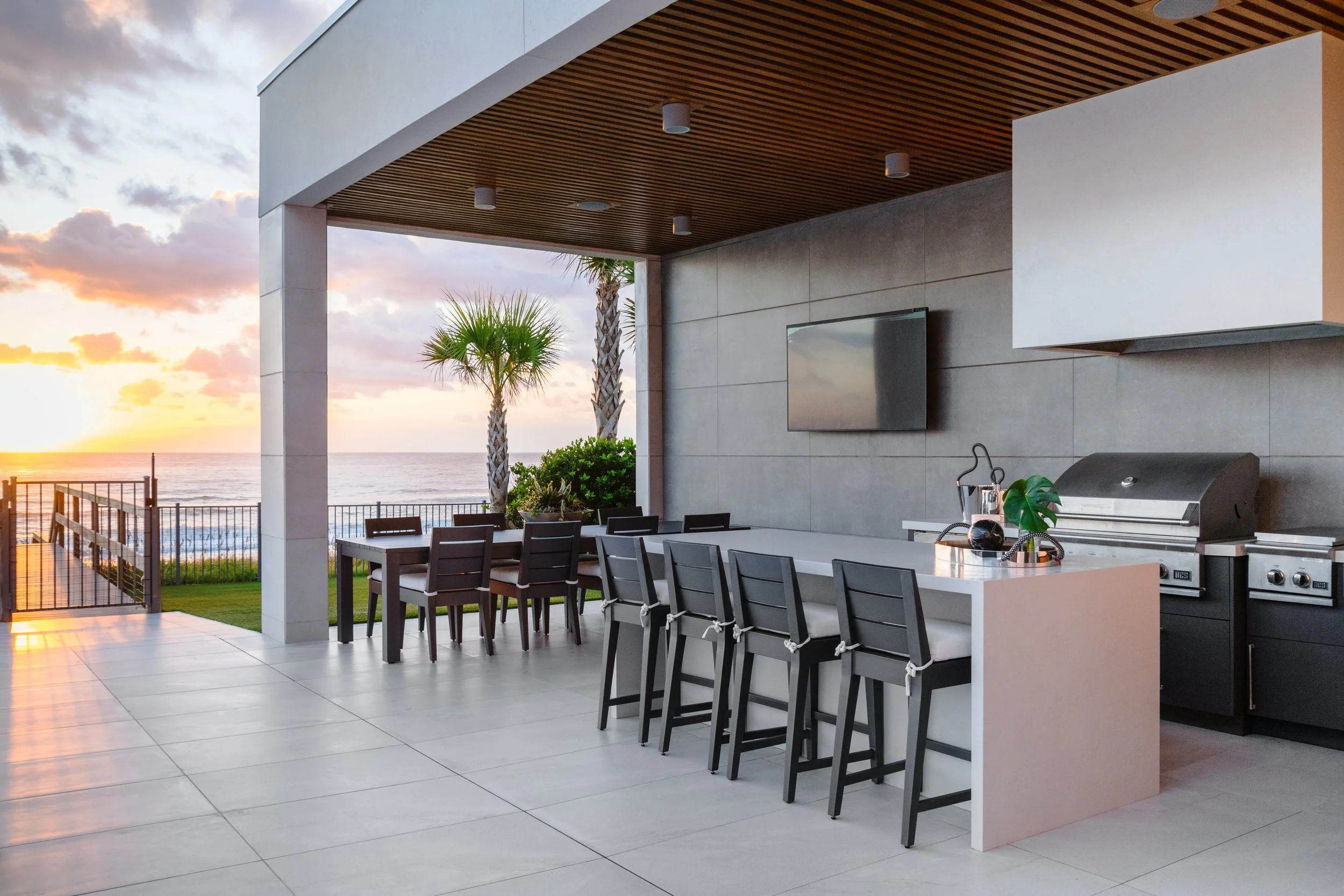 Outdoor patio area at sunset with a view of the ocean, featuring a dining table with chairs, a grill, potted plants, and a wall-mounted TV.