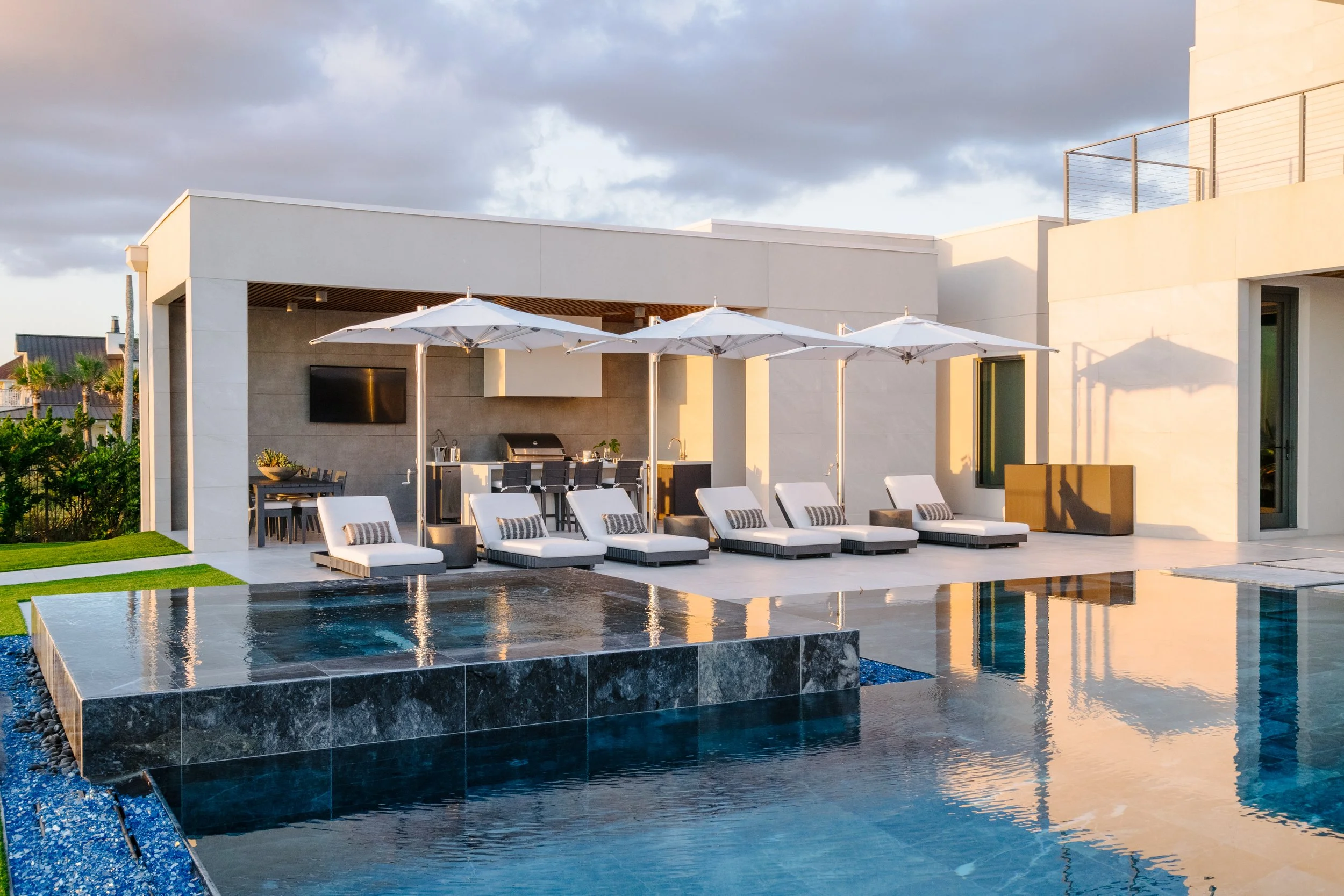 Modern outdoor patio with a swimming pool, sun loungers with striped cushions, white umbrellas, and a built-in outdoor kitchen area, illuminated by warm evening sunlight.