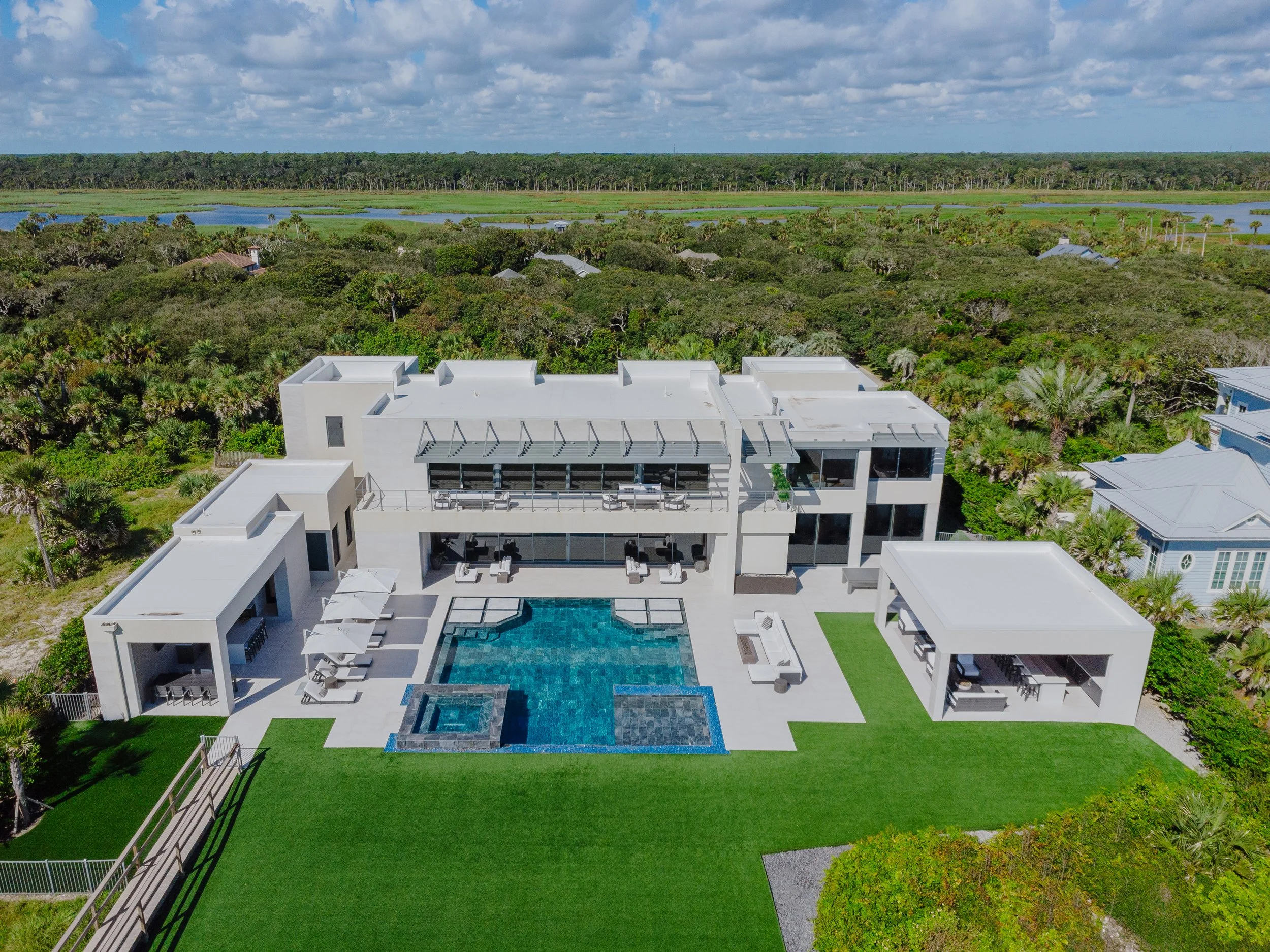  aerial view of a modern white house with a swimming pool surrounded by a green lawn, with lush trees and a natural landscape in the background under a partly cloudy sky