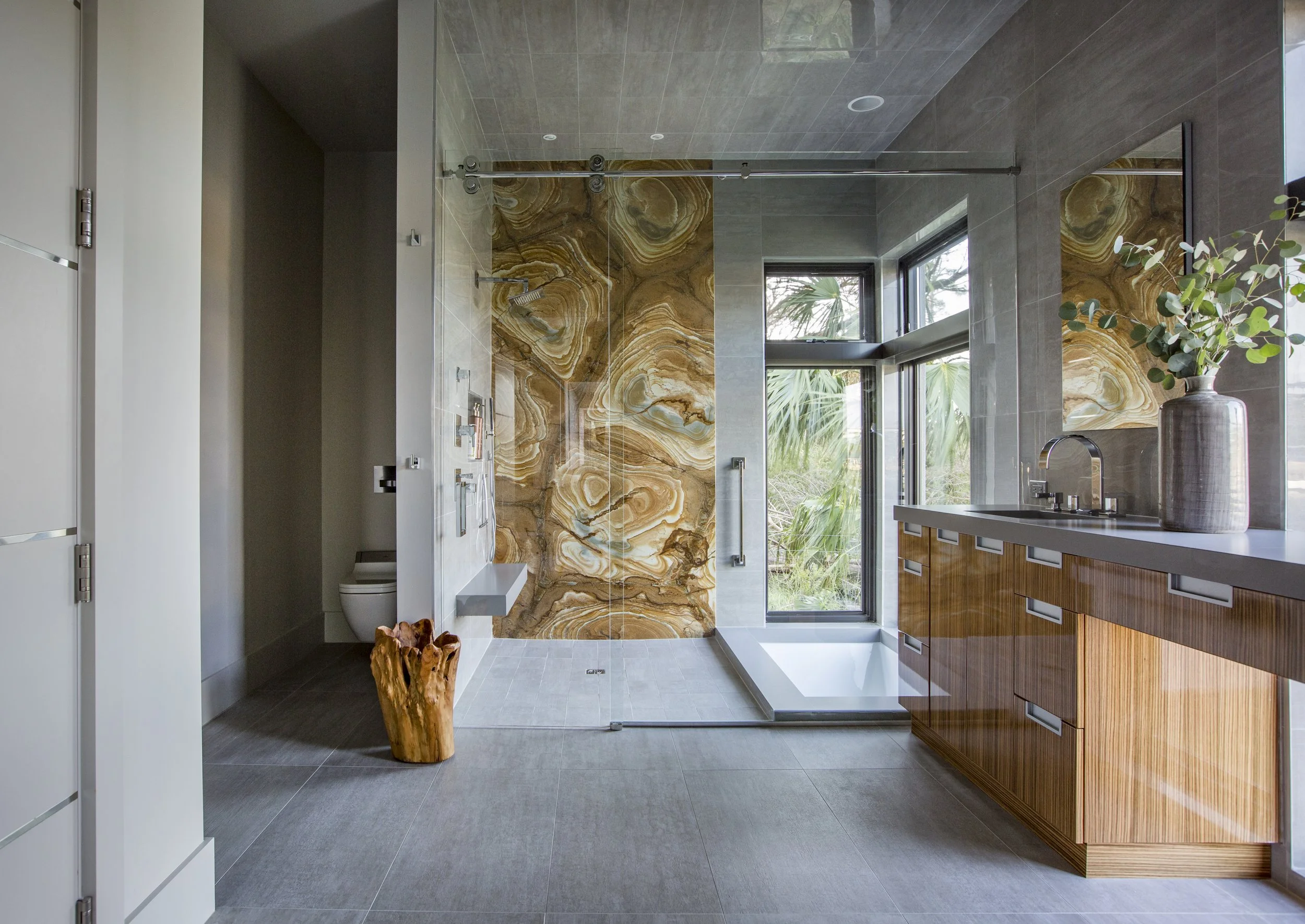 Modern bathroom with gray tiled floor and ceiling, large window with view of greenery, wooden vanity with drawers, and a large decorative stone wall in the shower area, which has glass doors.
