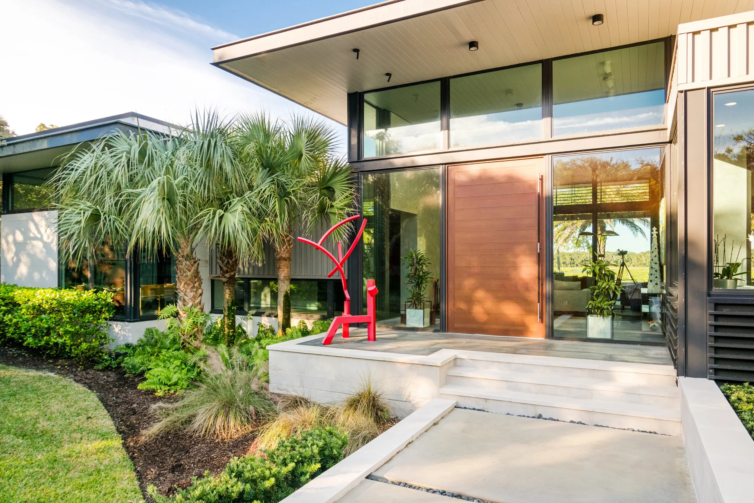 Modern house exterior with large glass windows, wooden door, steps, green plants, palm trees, and a clear sky.