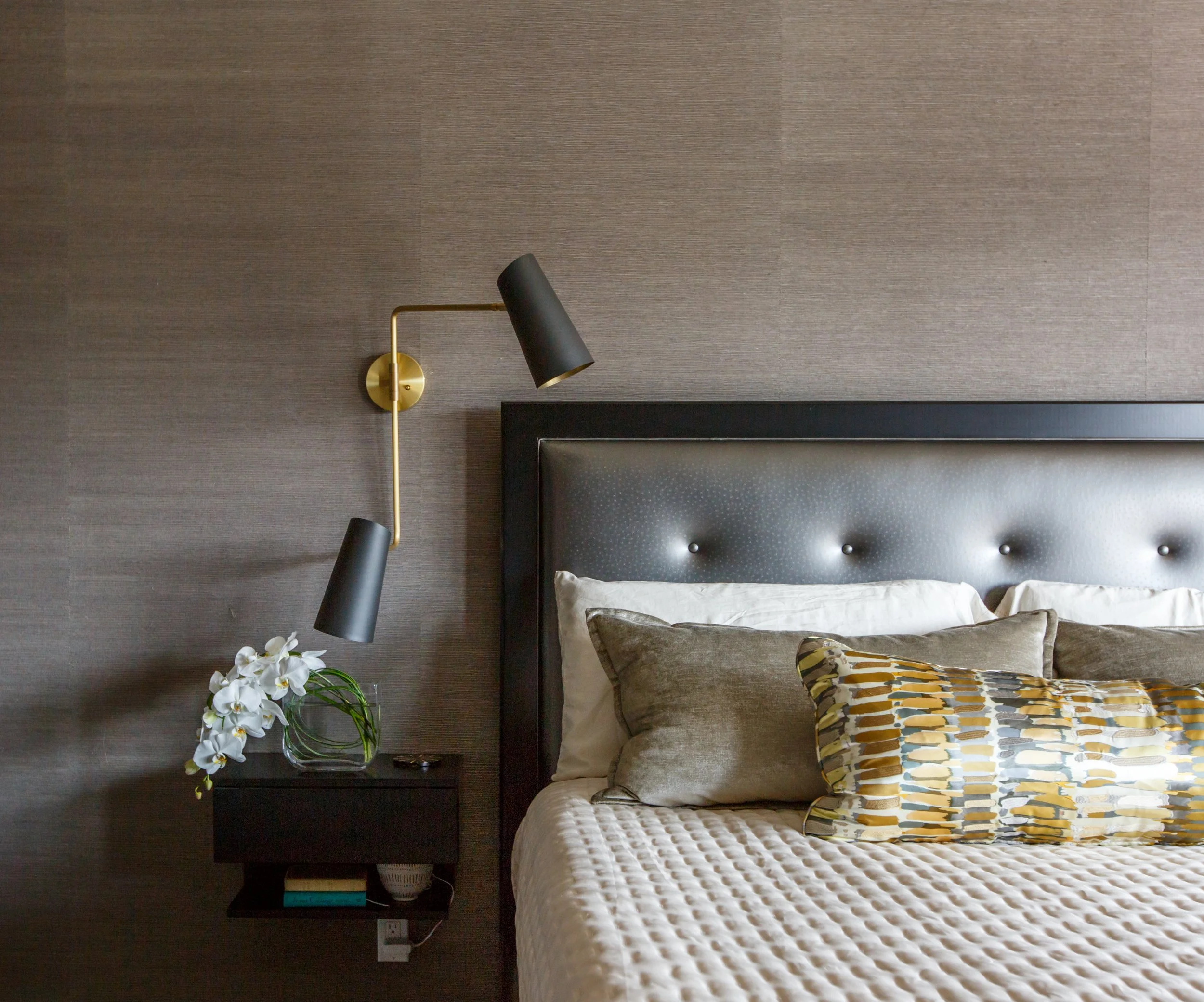 A bedroom with a bed that has a cream-colored quilted bedspread and a decorative pillow with a yellow, gray, and white pattern. To the left of the bed is a black nightstand with a glass vase containing white orchids. The wall behind the bed is covere