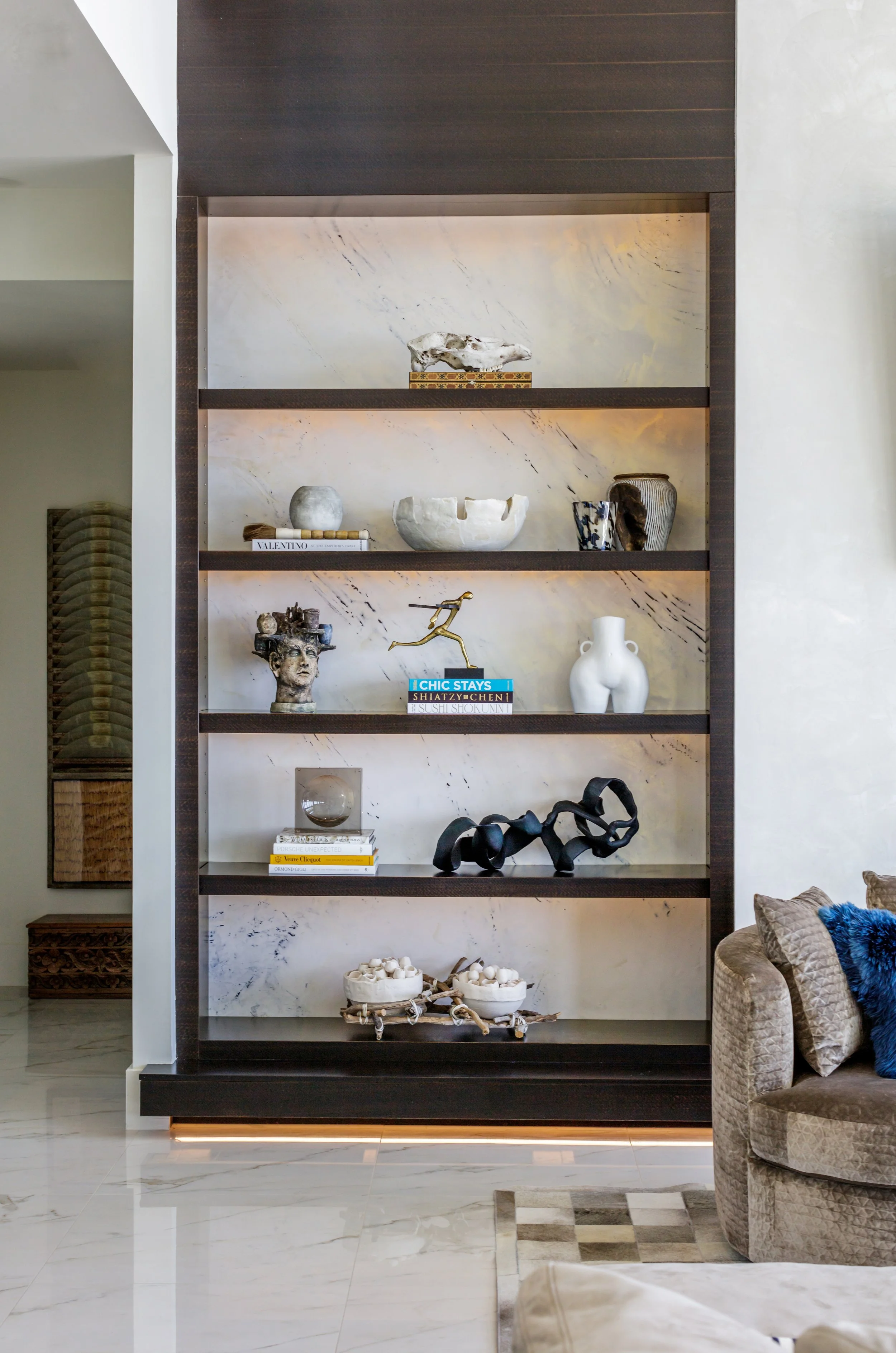 A decorative black bookshelf with gold trim, displaying sculptures, books, and vases against a marble wall. To the right, a beige patterned armchair with blue and beige pillows is partially visible.