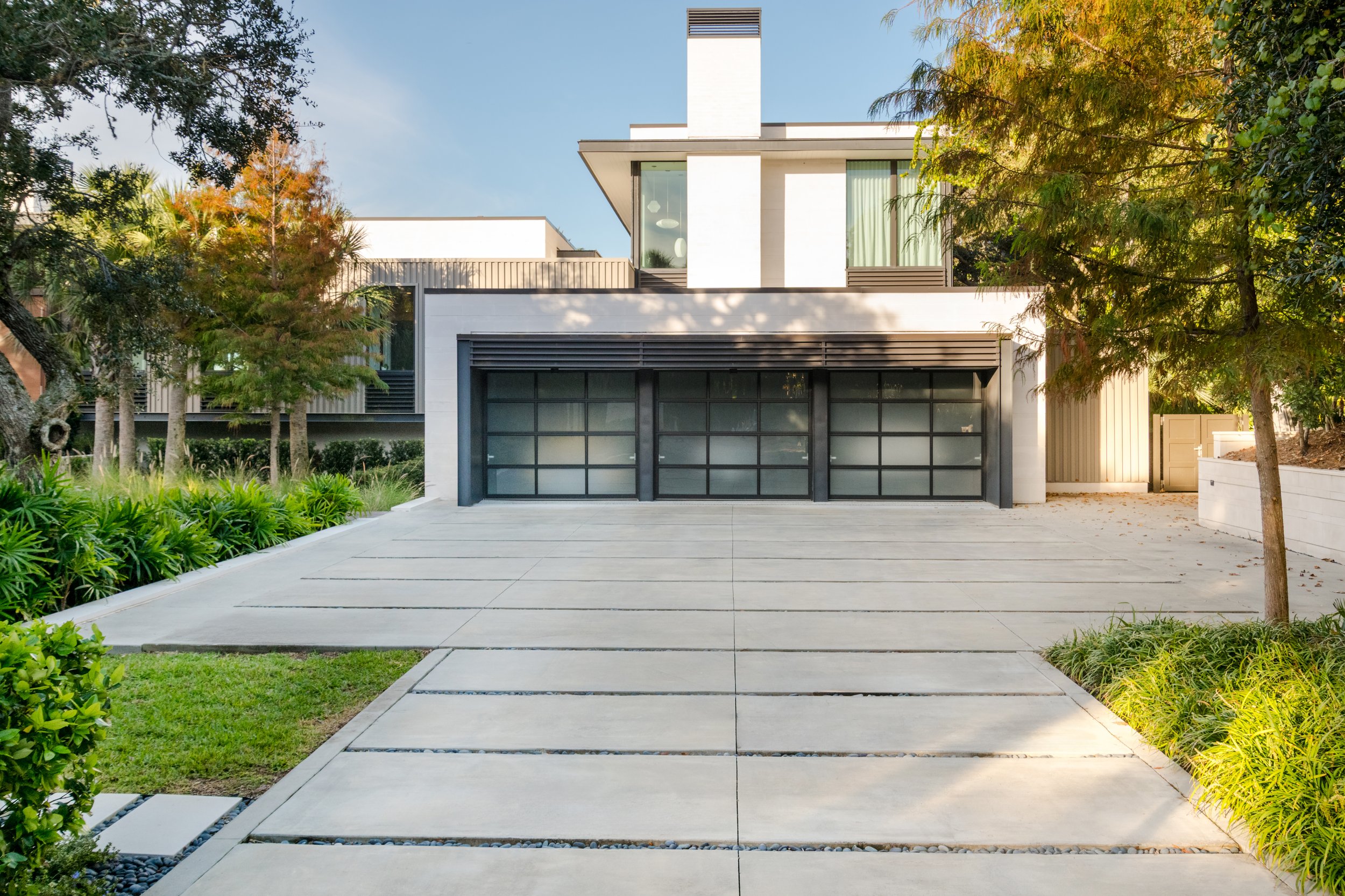 Modern white house with large black garage doors, surrounded by lush greenery and trees, with a paved driveway and pathway leading to the front door, under a clear blue sky.