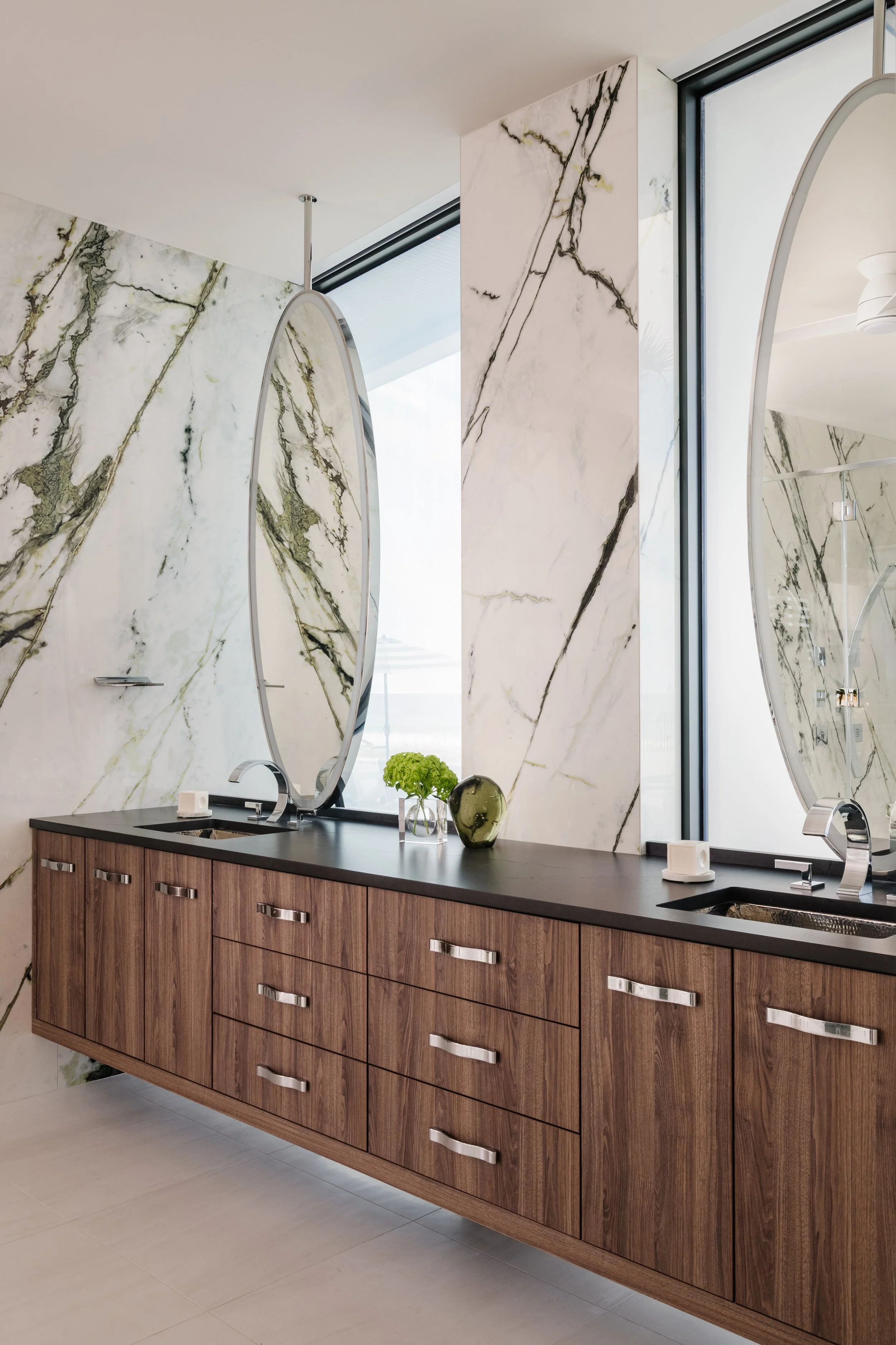Bathroom vanity with wooden drawers, black countertop, oval mirror, faucet, decorative green plant, and a window with marble wall behind.