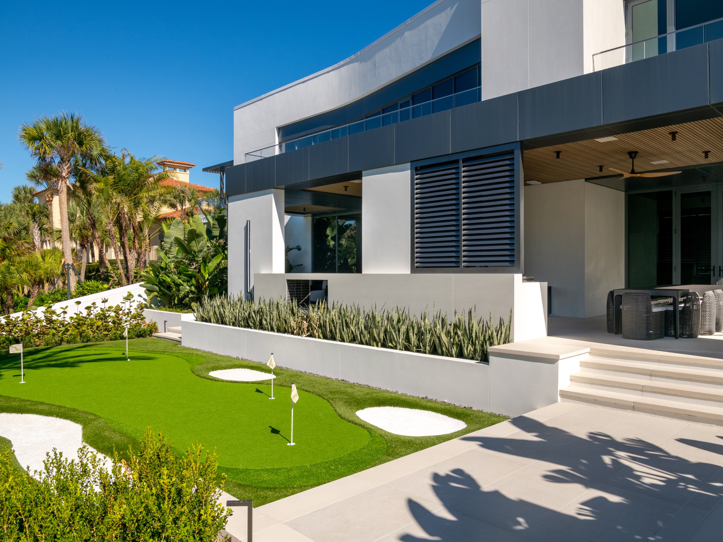 Modern white residential building with glass balcony, surrounded by lush green plants, palm trees, and a golf putting green with flags in the foreground under a clear blue sky.
