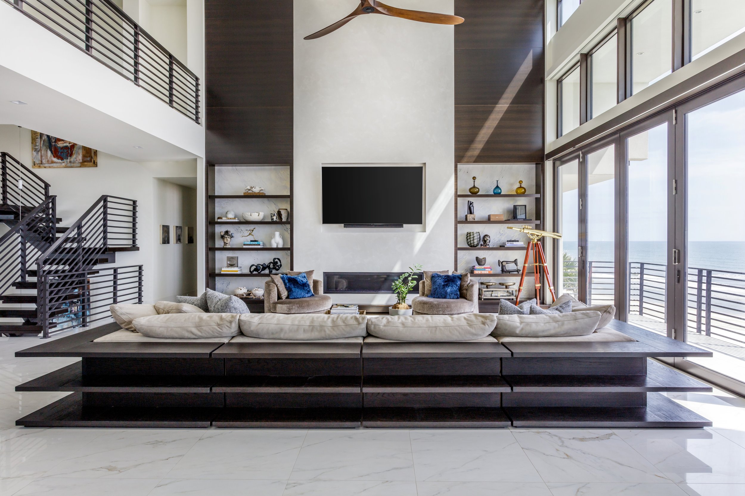 Modern living room with large windows, a wall-mounted flat-screen TV, built-in shelves with decorative items, beige sectional sofa with blue pillows, and a view of the ocean outside.