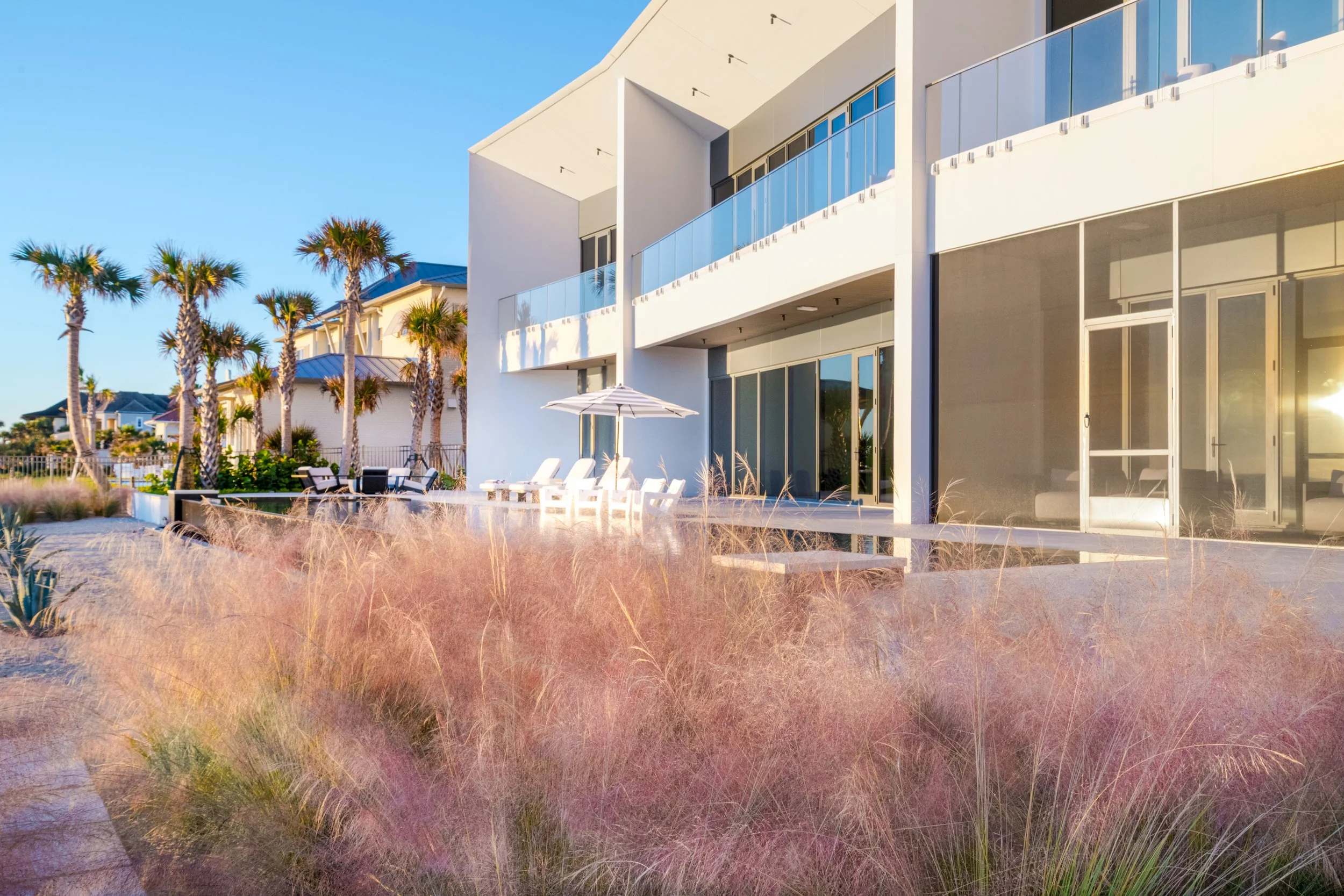 Modern white beach house with large glass windows and a balcony, surrounded by palm trees and beach chairs under umbrellas, with a sandy ground and ornamental grasses in the foreground under clear blue sky.