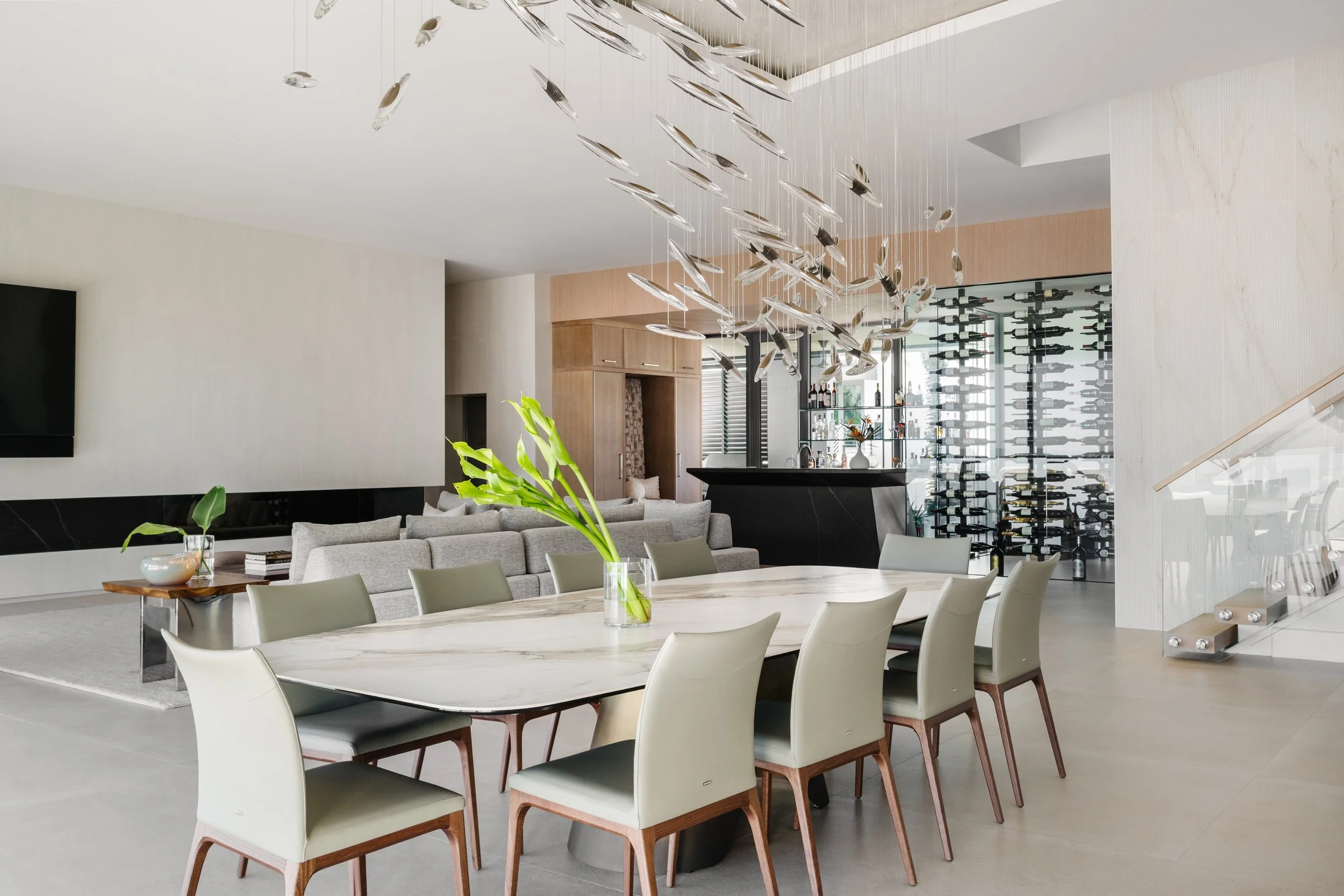 Modern open-concept living and dining area with a large marble dining table, white chairs, a gray sofa, and a wine wall with bottles. Central hanging light fixture with multiple glass elements. Plants add a touch of greenery.