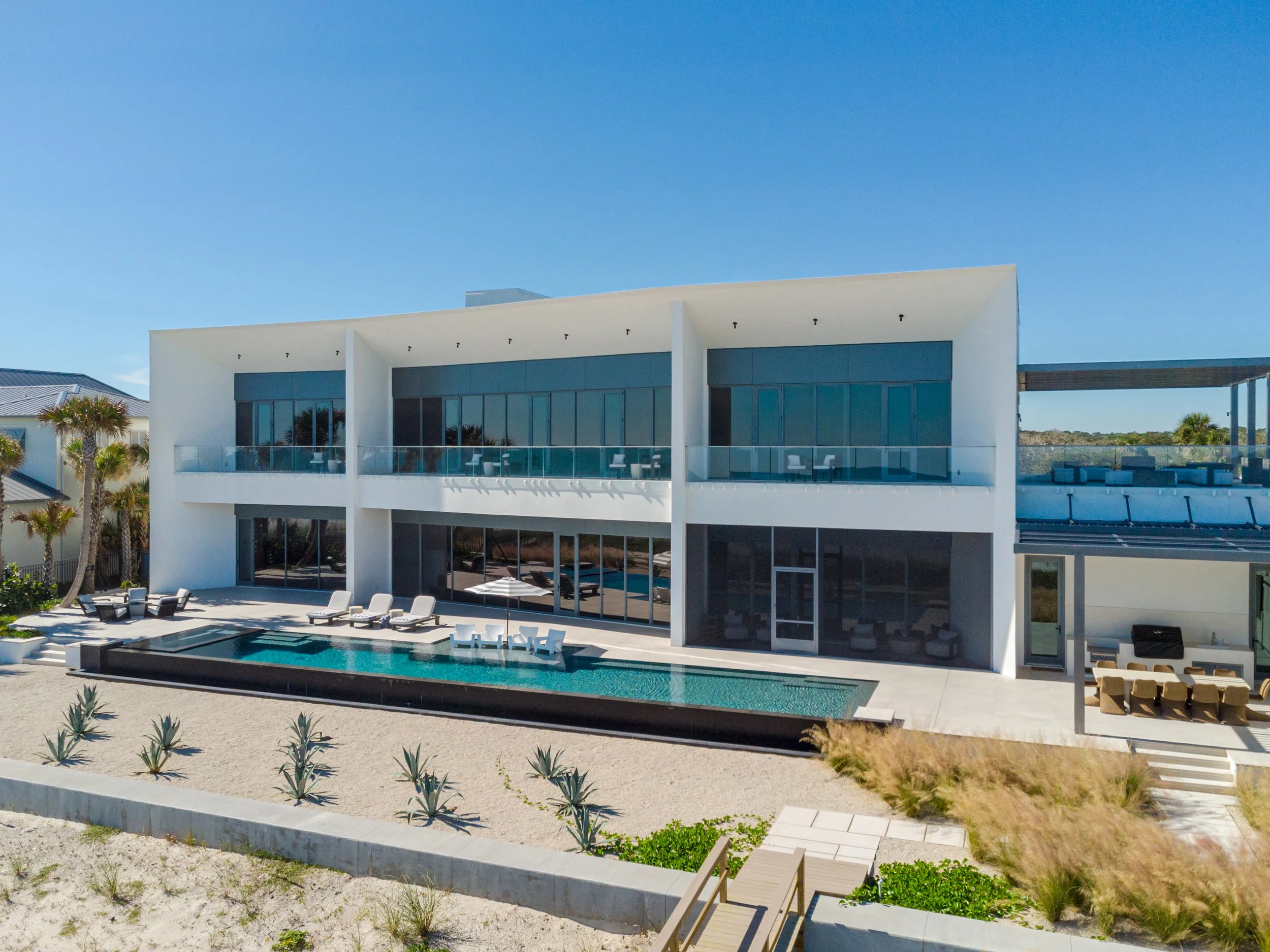 Modern white house with large glass windows and a swimming pool in the backyard, surrounded by lounge chairs and palm trees.
