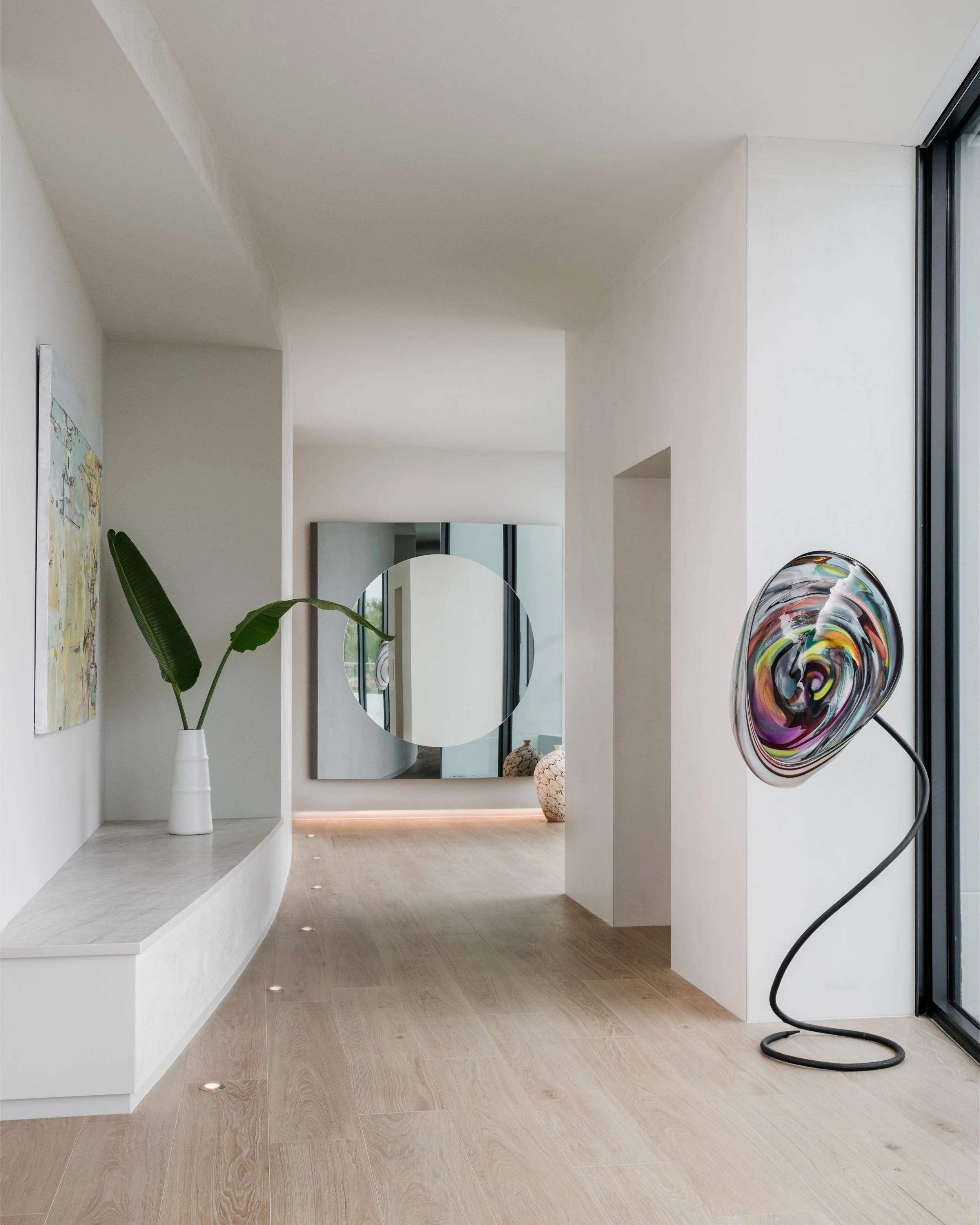 Modern interior space with light wood flooring, white walls, a white built-in ledge with small recessed lighting, a tall white vase with green leaves, a mirror with rounded edges, a colorful glass sculpture on a black stand, and artwork on the wall.