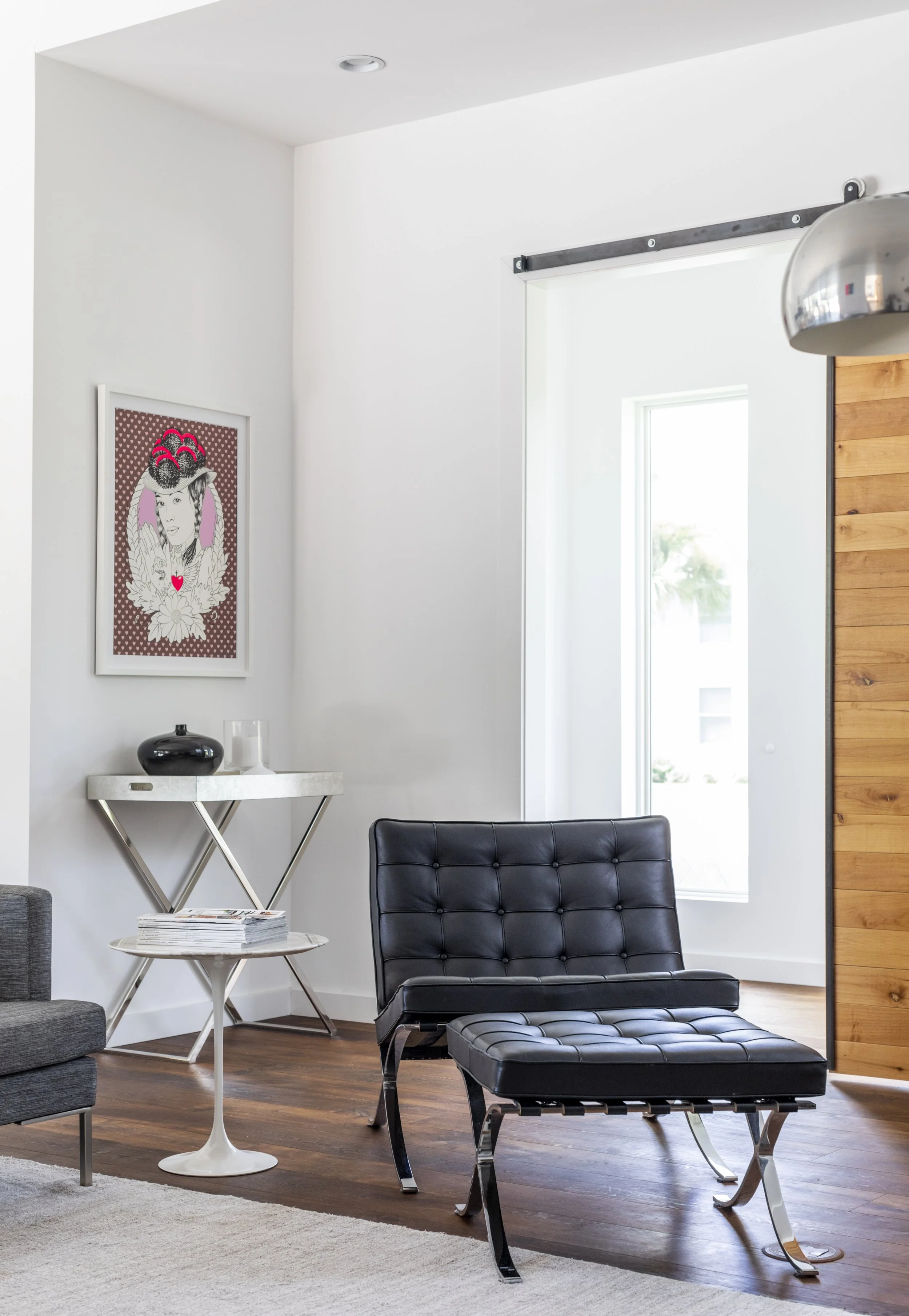 Modern living room with black leather lounge chair and ottoman, white side table with magazines, abstract art on the wall, and a wooden accent wall near a large window.