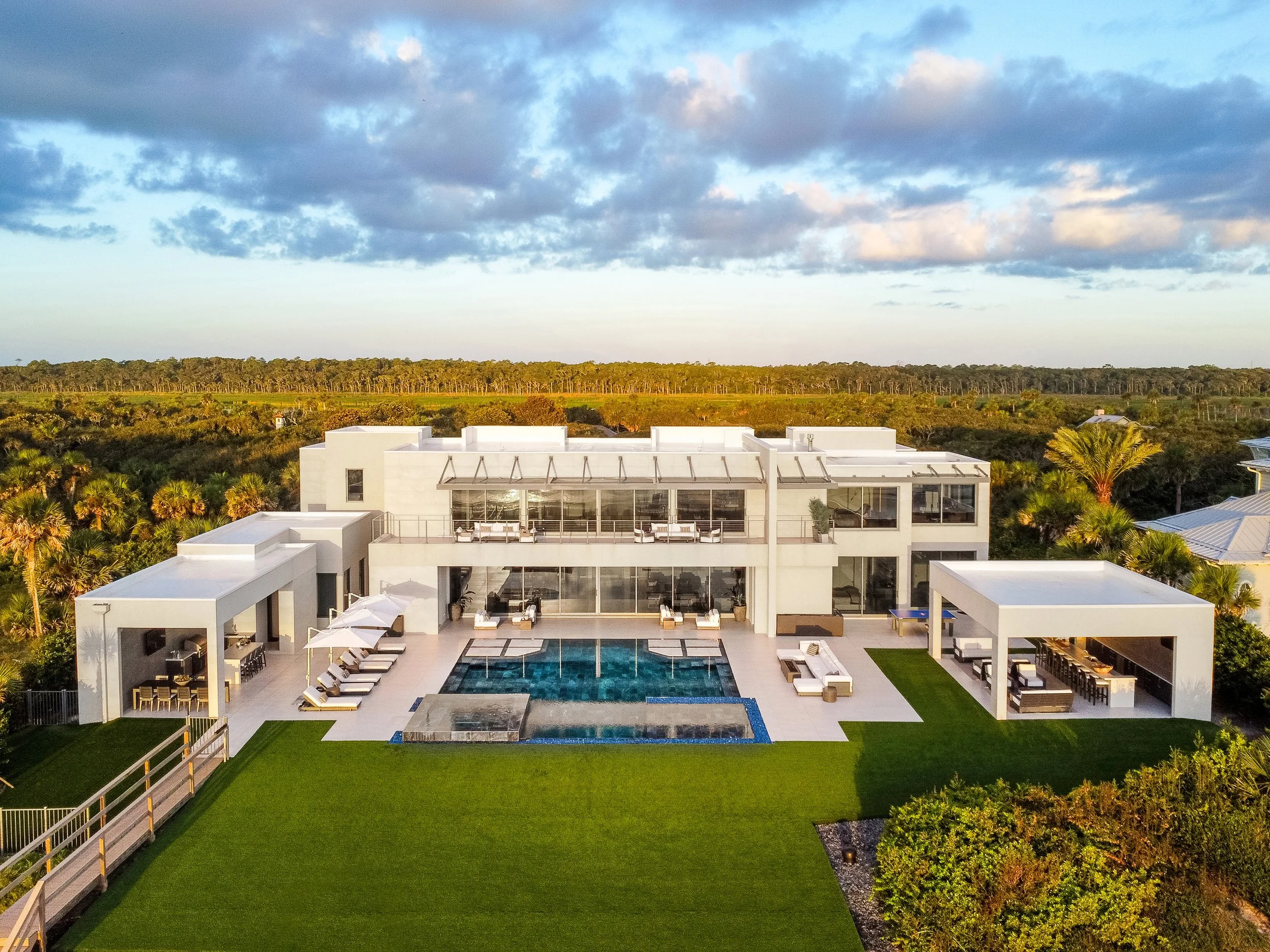 A modern white house with an outdoor pool and patio area, surrounded by a green lawn, trees, and a forested landscape under a partly cloudy sky.