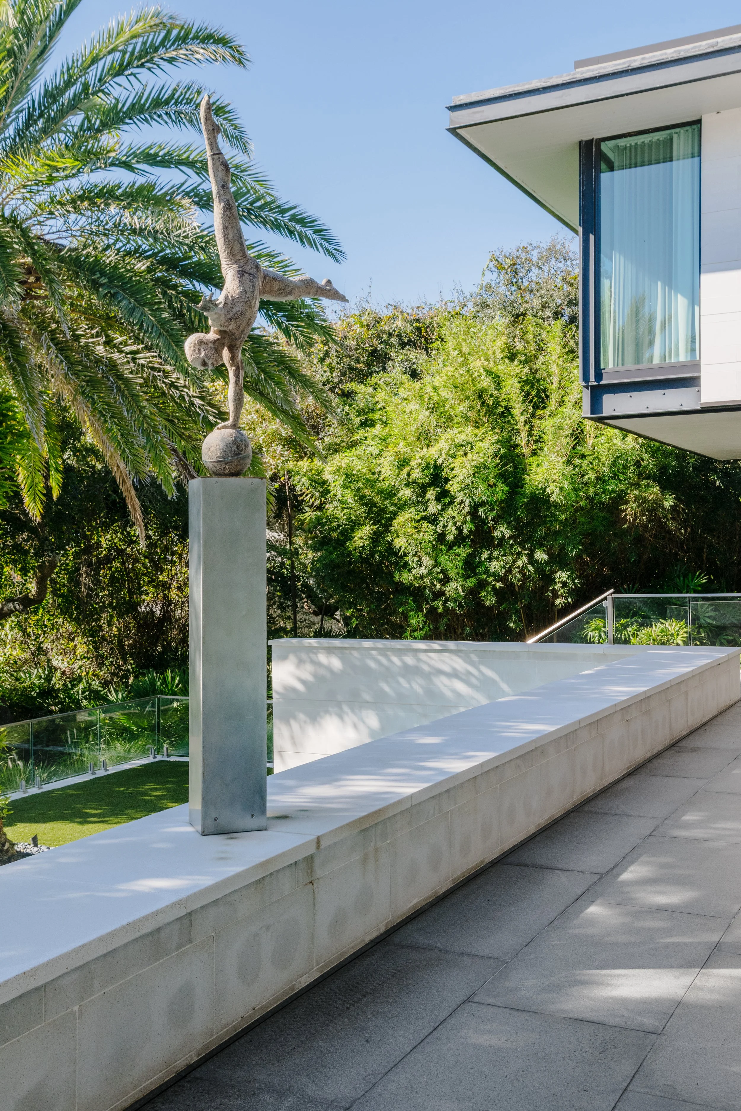 Modern white building with a glass balcony, surrounded by lush green trees and a sculpture of a person on a ball on a tall pedestal, clear blue sky.