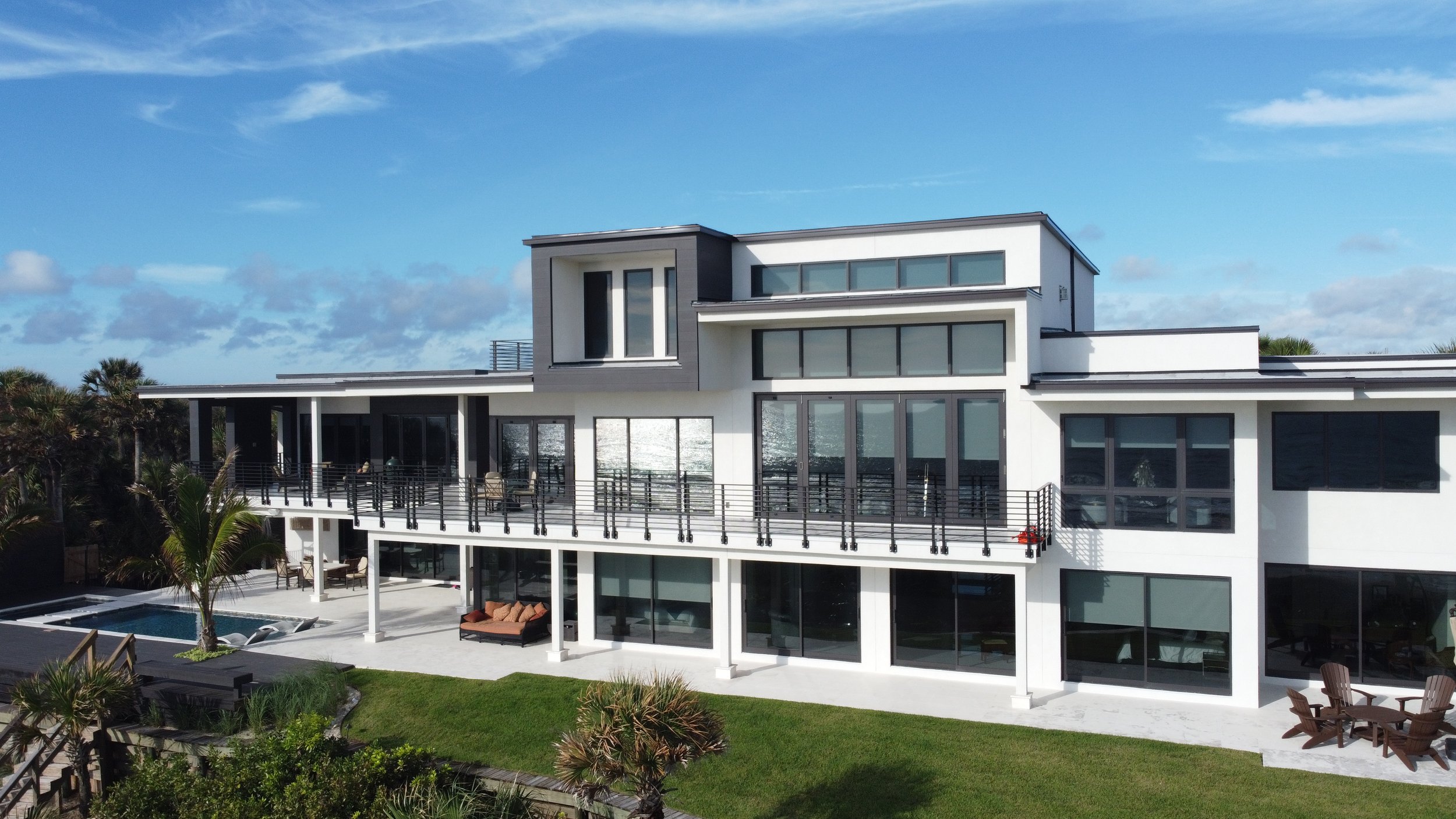 Modern multi-story house with large glass windows, balcony, outdoor seating, lush green lawn, swimming pool, and palm trees against a blue sky with clouds.