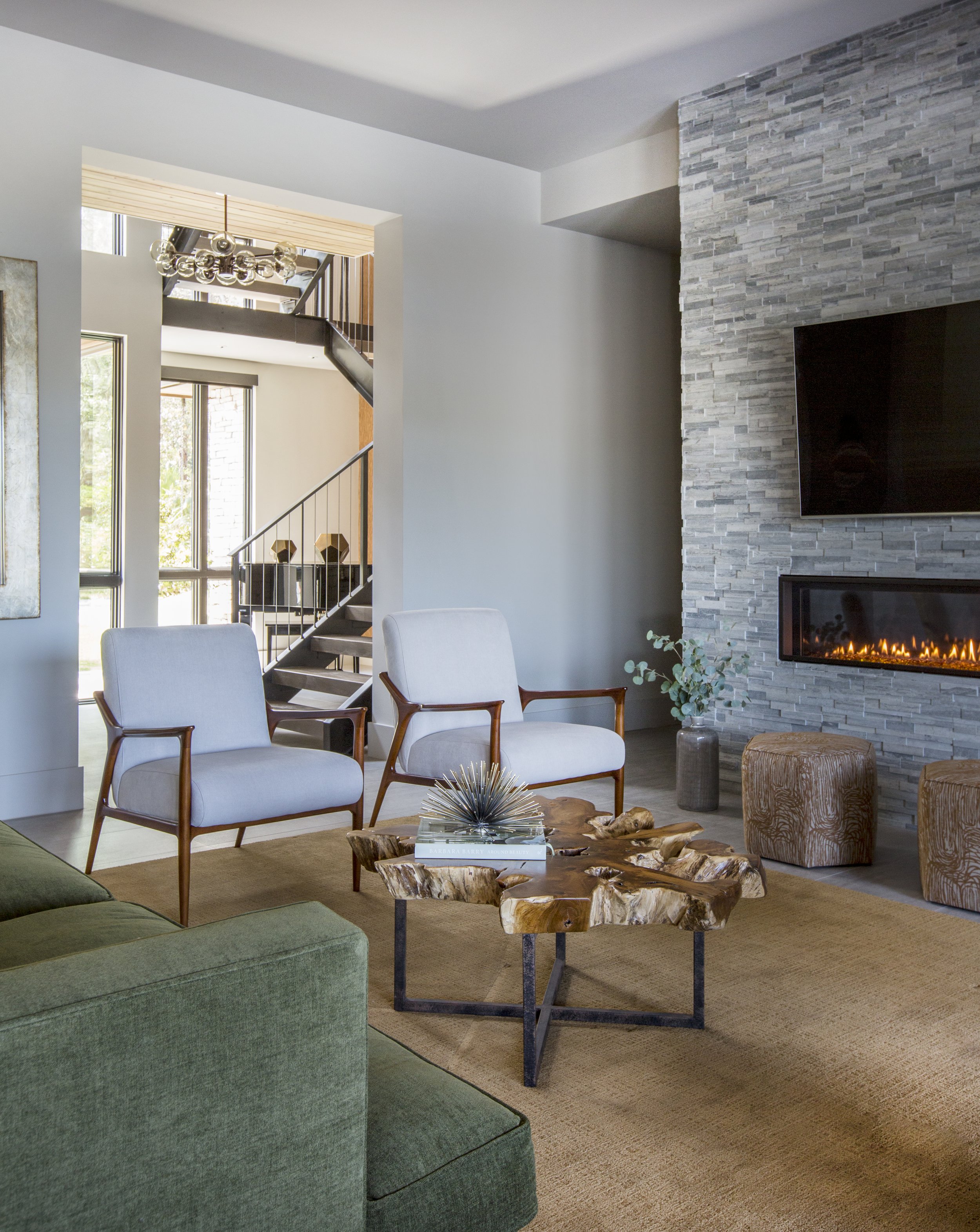 Living room with green sofa, two white armchairs, a wooden coffee table, and a stone fireplace with a fire. Large windows, a staircase visible in the background, and decorative items on the table and floor.