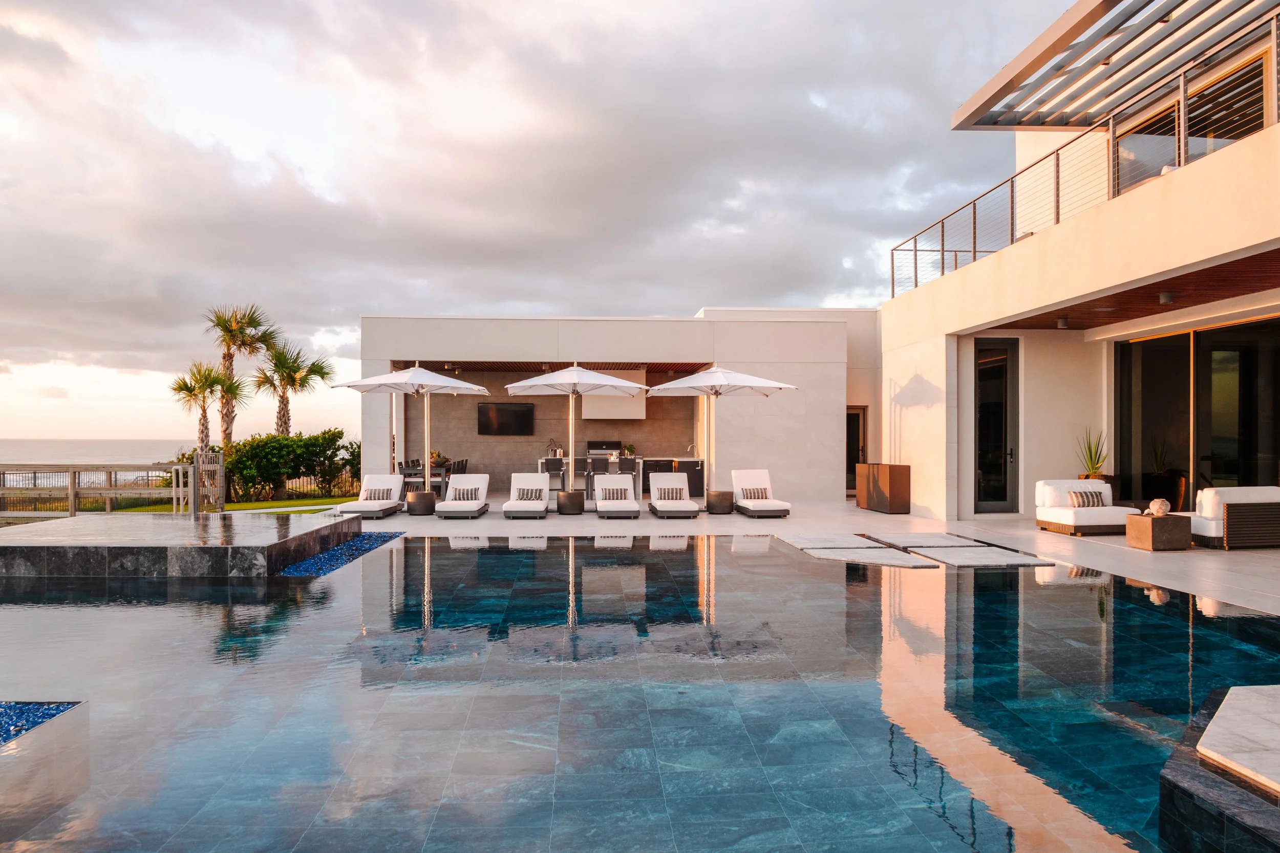 Modern house with outdoor pool, sun loungers, umbrellas, and palm trees, overlooking the ocean during sunset.