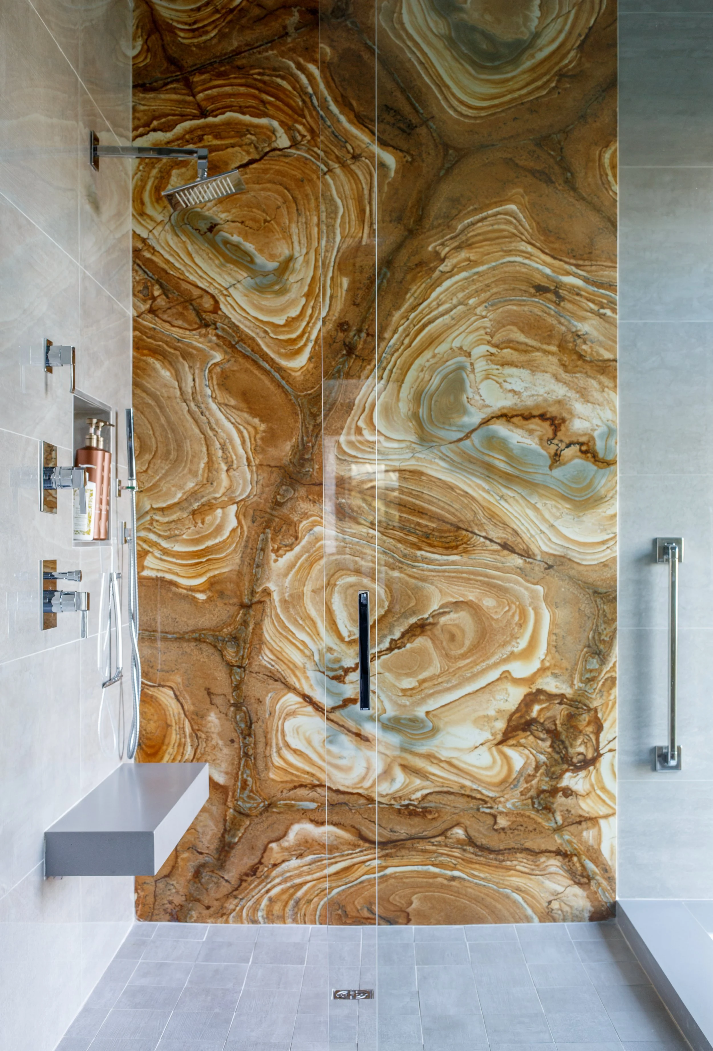 A modern bathroom shower with a striking marble wall featuring swirling brown, white, and tan patterns. The shower has a glass door, a built-in shelf, a metallic showerhead, and a handheld shower wand, with light gray tiled flooring.