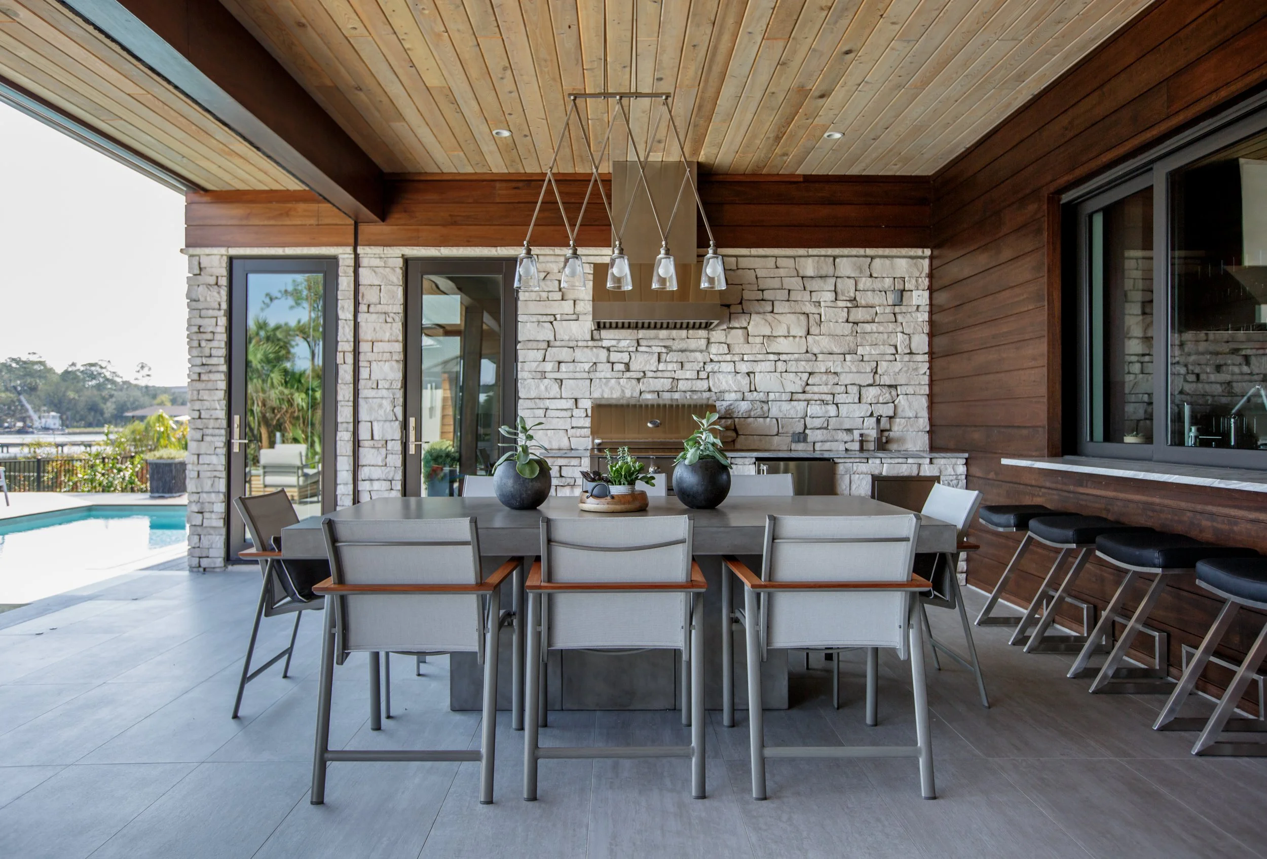 Modern outdoor dining area with a large table, surrounded by chairs, under a wood-paneled ceiling. The space features stone and wooden wall accents, with a chandelier hanging above the table. Large sliding glass doors open to a patio and pool area wi