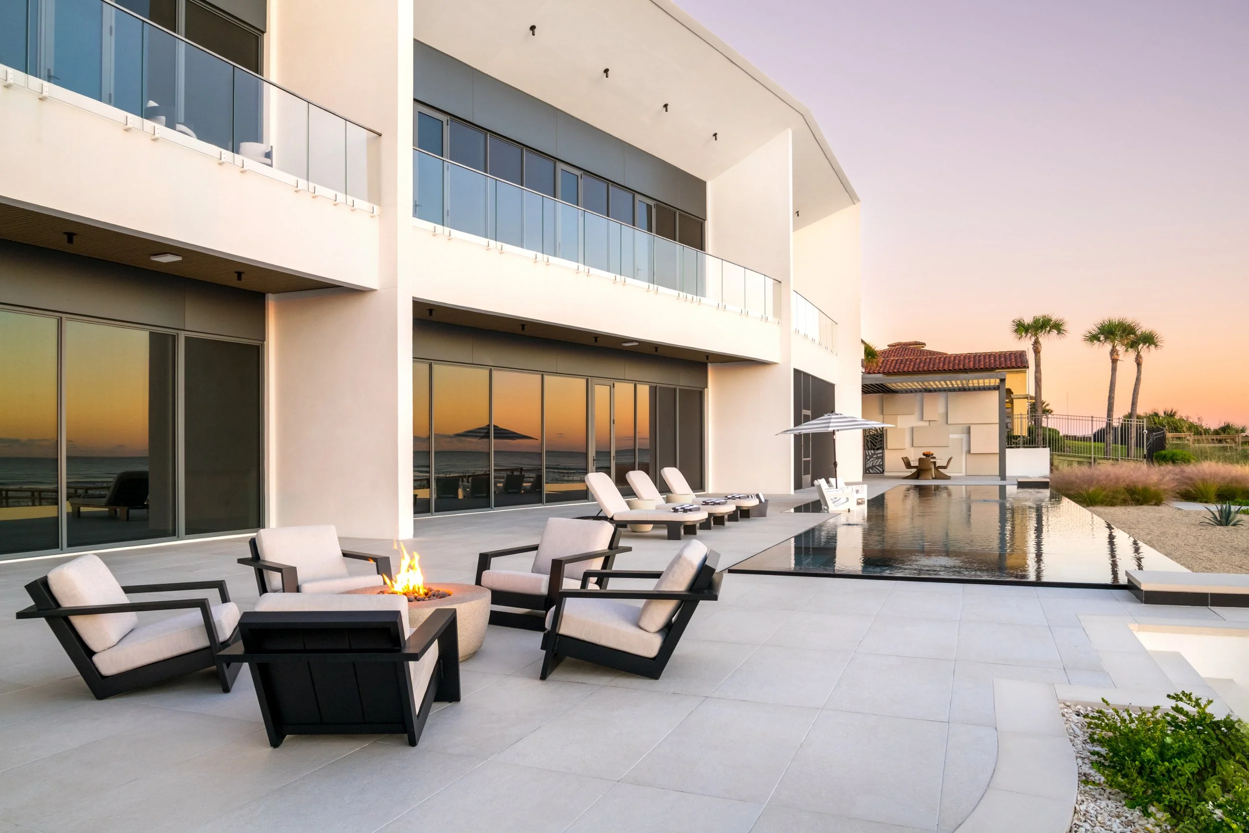 Modern white house with large glass sliding doors and a balcony, overlooking a patio with lounge chairs, an outdoor fire pit, and a rectangular pool at sunset, with palm trees and a fence in the background.