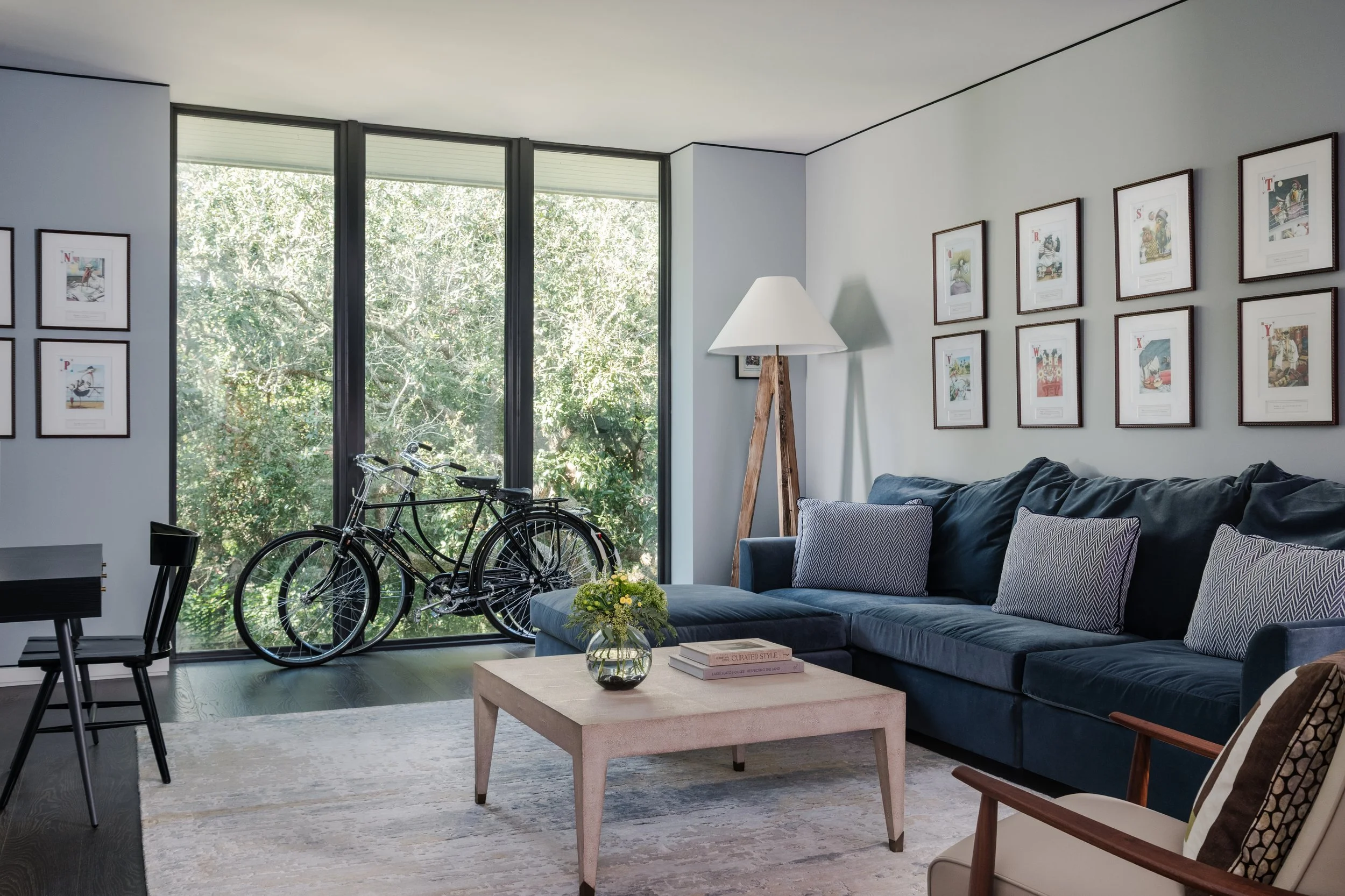 Modern living room with a large sectional sofa, framed artwork on the wall, a wooden floor lamp, a glass coffee table with books and a flower arrangement, a bicycle near large window, and outdoor greenery visible through the window.