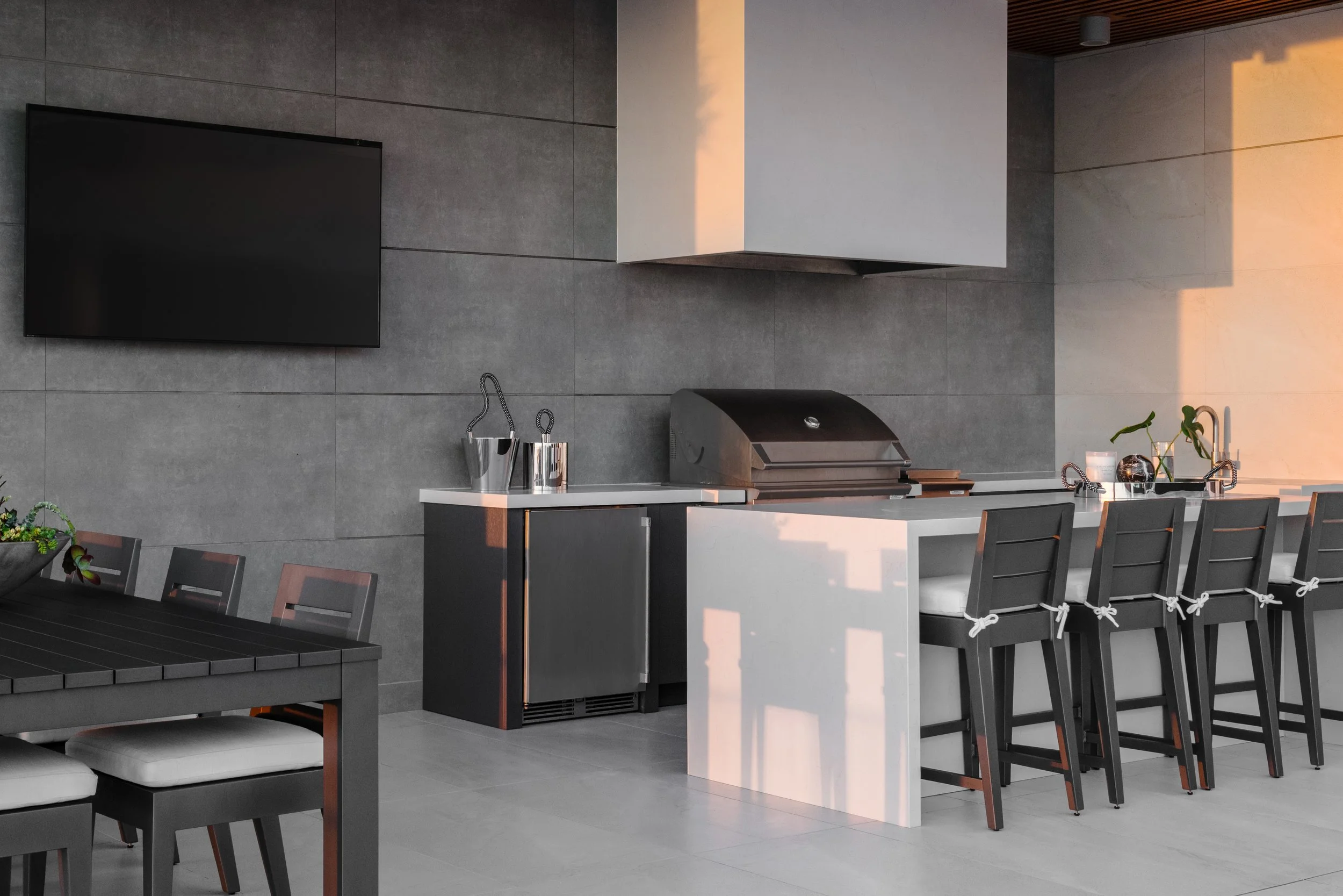 Modern kitchen with gray tiled walls, white countertop, black dining chairs, a black television mounted on the wall, and a barbecue grill.
