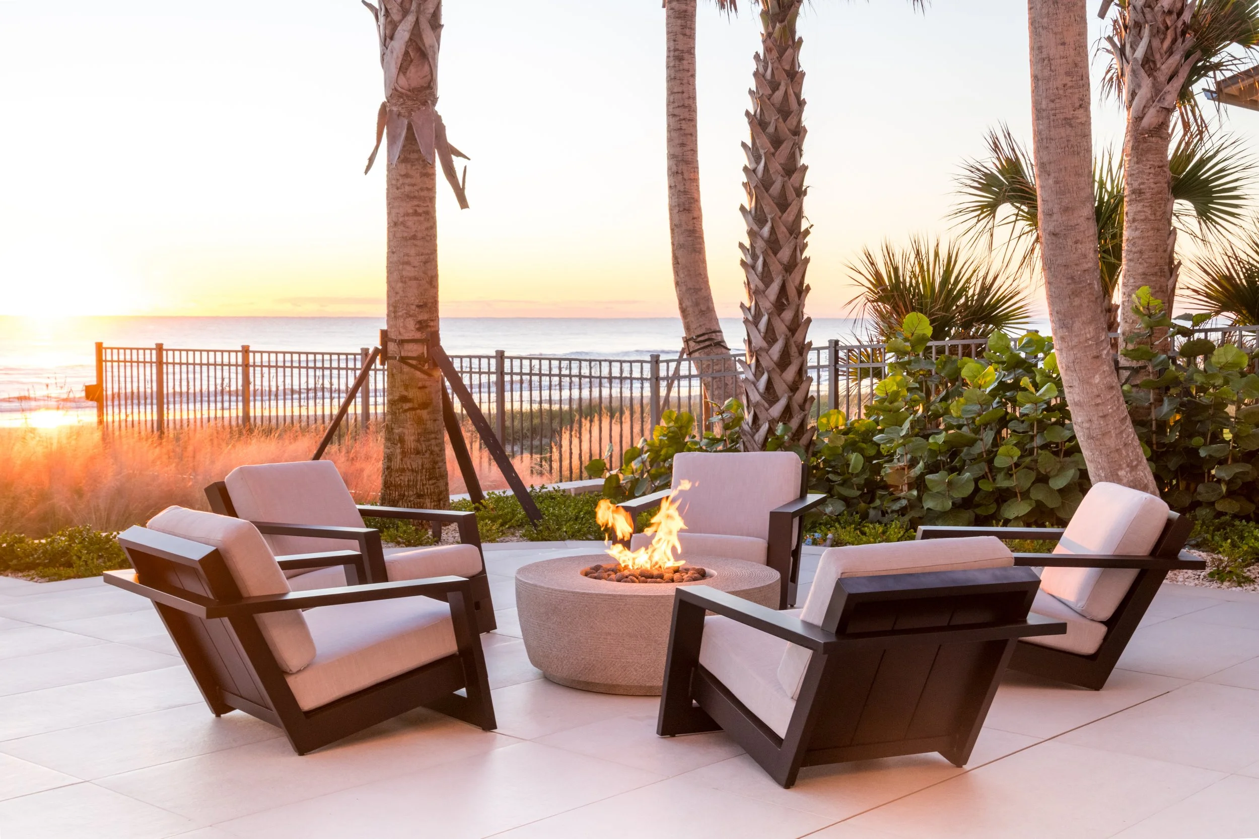 Outdoor seating area with six cushioned chairs arranged around a fire pit, overlooking the beach at sunset. Tall palm trees line the background with a fence separating the area from the beach, and the ocean is visible in the distance.