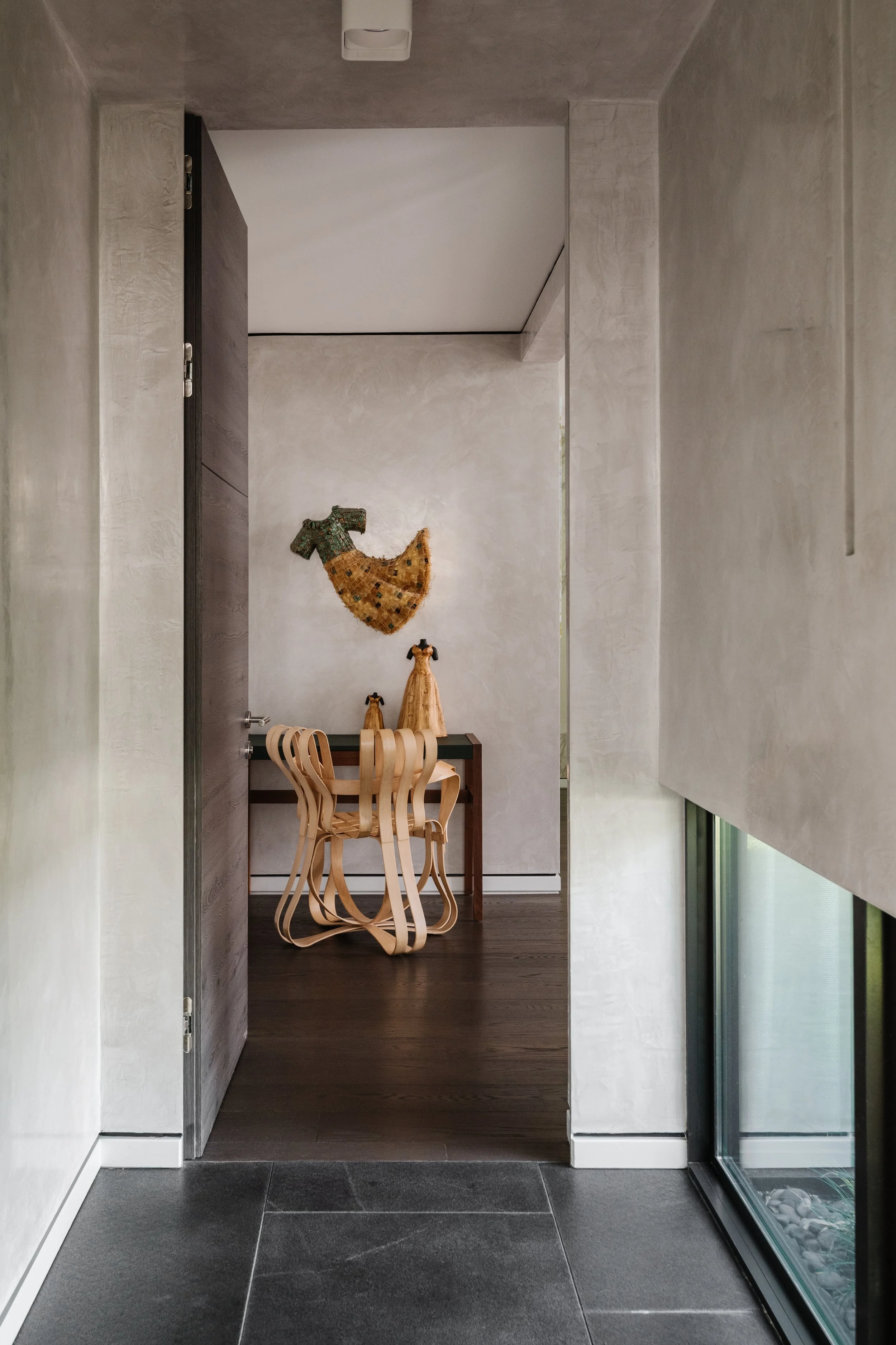 Modern interior space with a small desk, two dog sculptures, and a wall hanging shaped like a dog, in a room with wood flooring and a large window with a vertical blind.