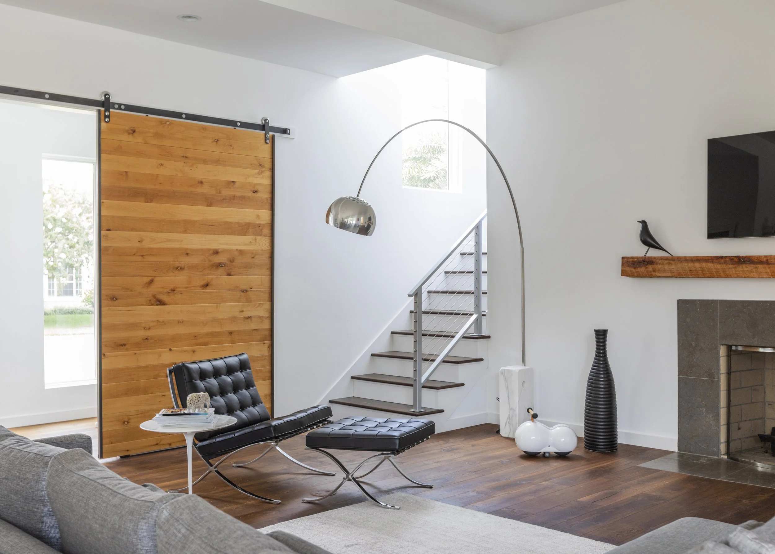 Living room with modern black leather chair and ottoman, gray sofa, wooden flooring, white walls, a staircase, a large flat-screen TV, and a decorative black vase, with a wooden sliding barn door and a large window in the background.
