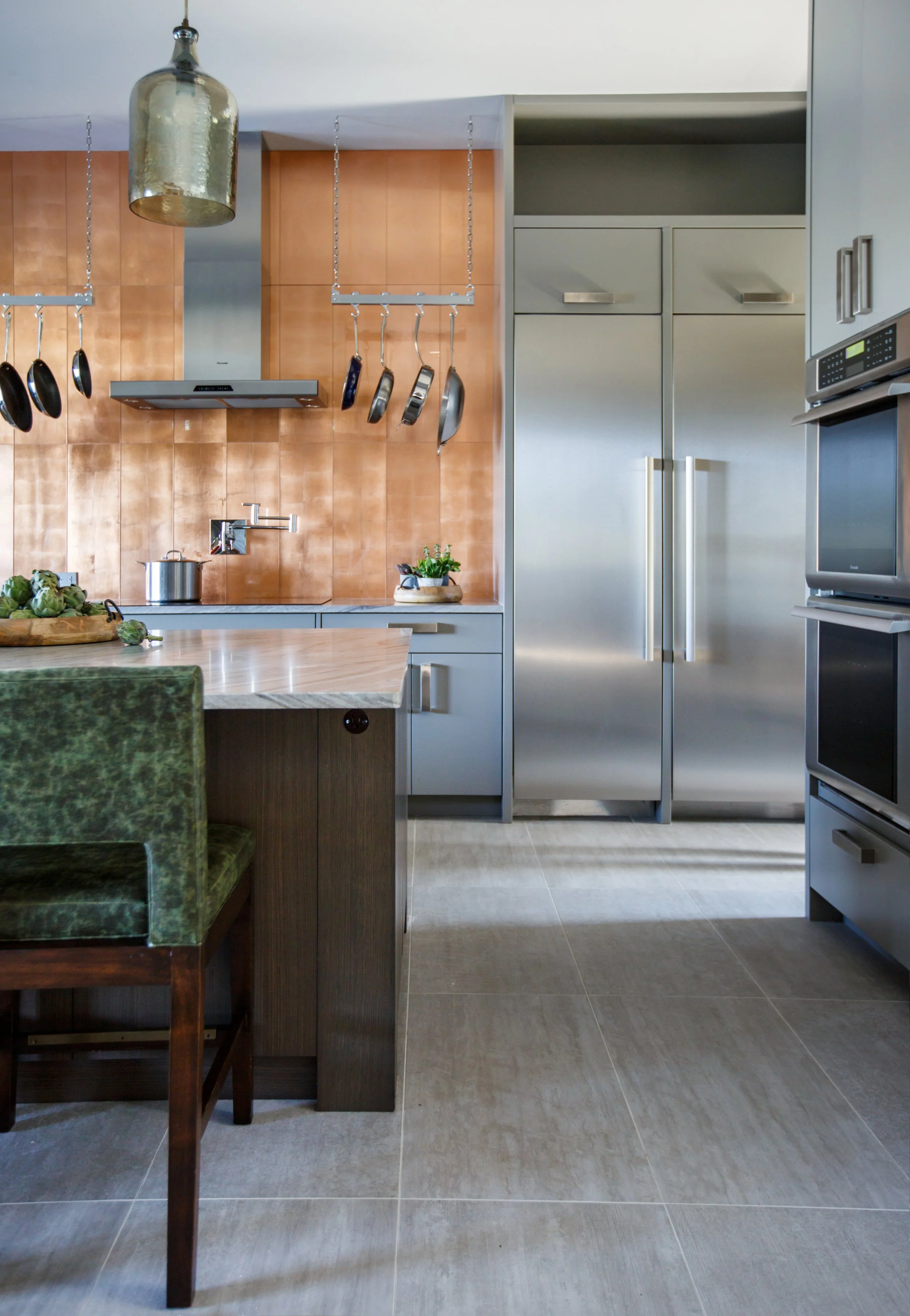 Modern kitchen with wooden backsplash, stainless steel refrigerator, hanging pots and pans, microwave, and a kitchen island with a green chair.