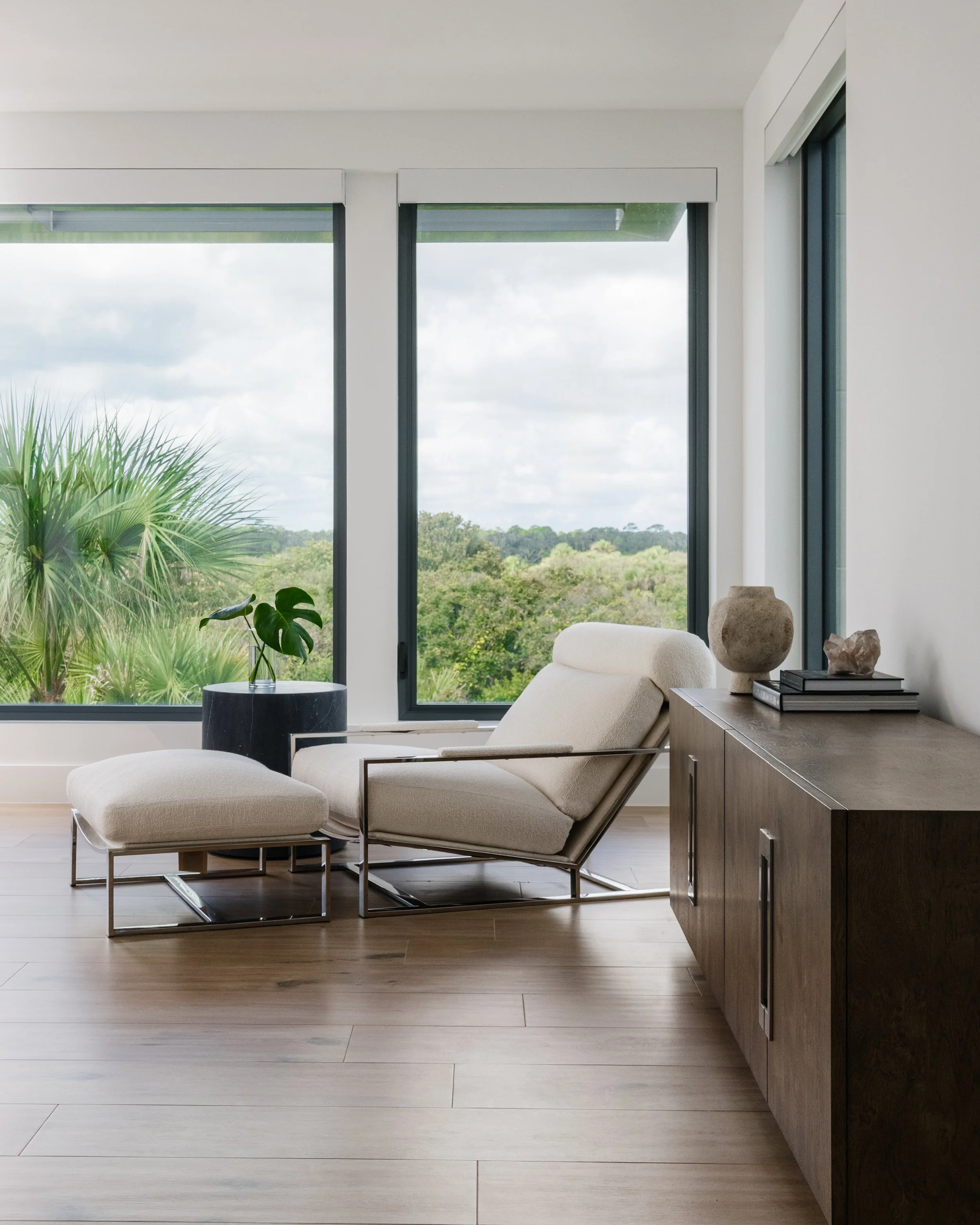 Living room with large windows showing green trees outside, a cream-colored armchair with matching footstool, a dark wooden sideboard with decorative stone objects, and a black side table with a potted plant.