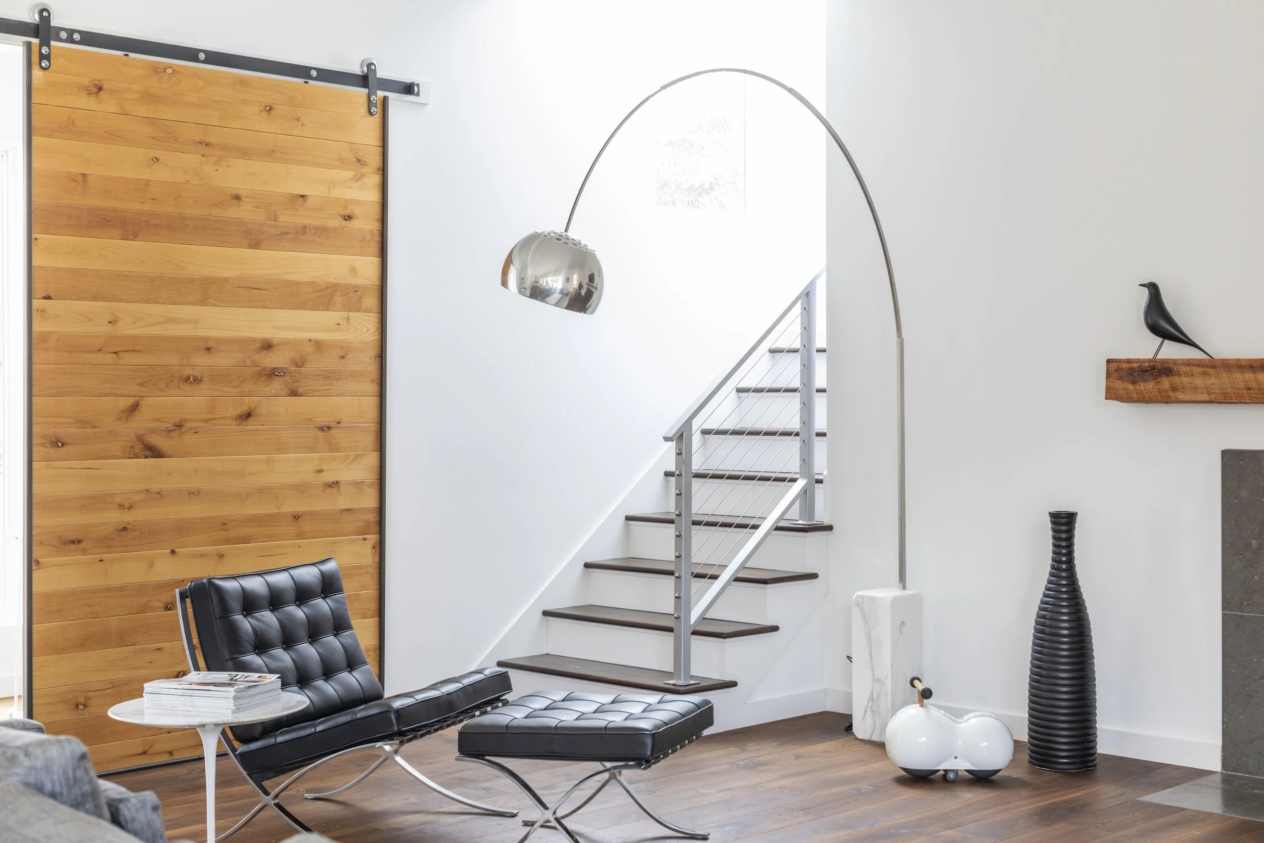 Modern living room with black leather furniture, a white coffee table, a wooden wall, a staircase with metal railing, a silver arc floor lamp, and decorative items including a black bird sculpture and a tall black vase.