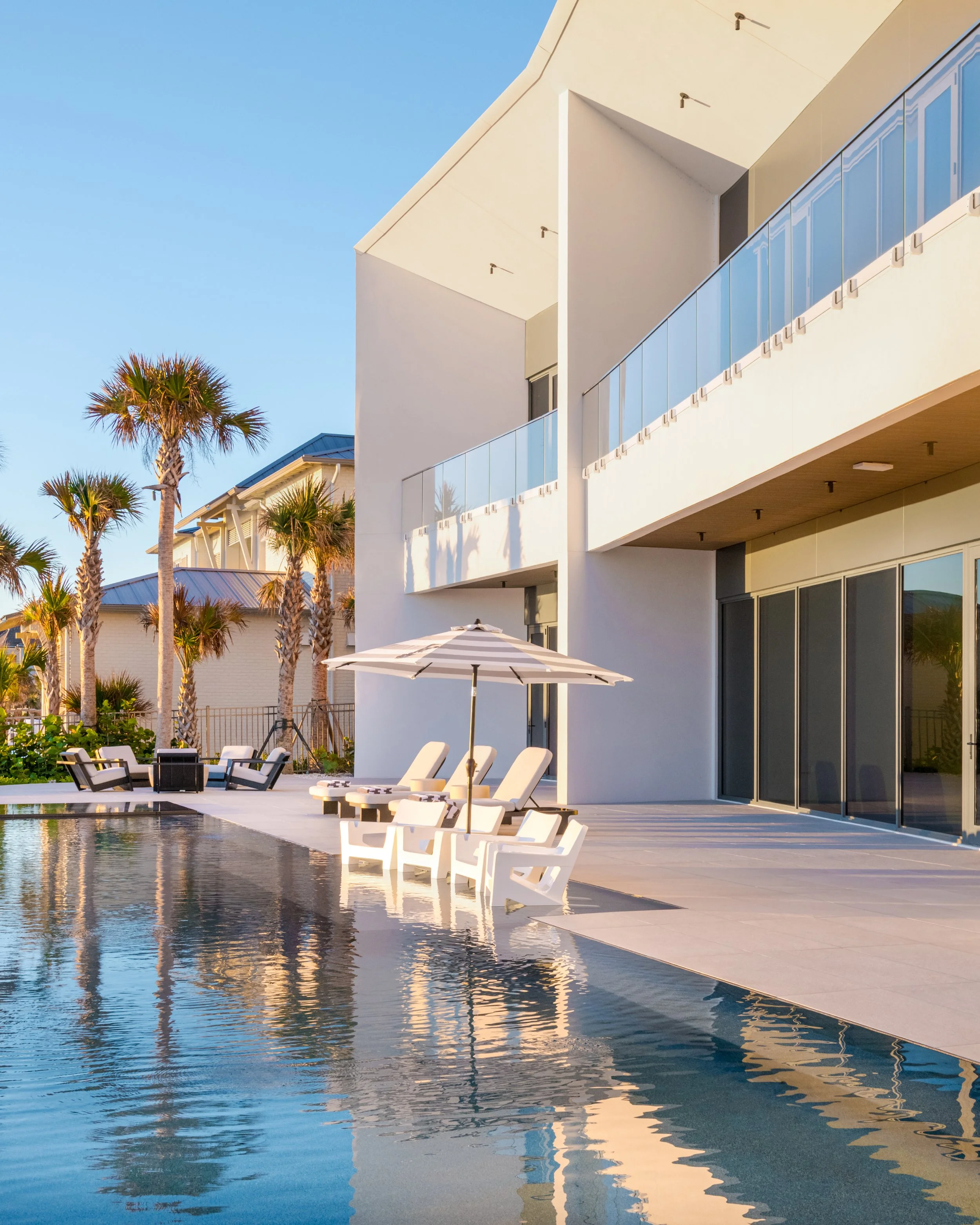 Modern white building with balconies, palm trees, outdoor seating, and a reflecting pool in a sunny, coastal setting.