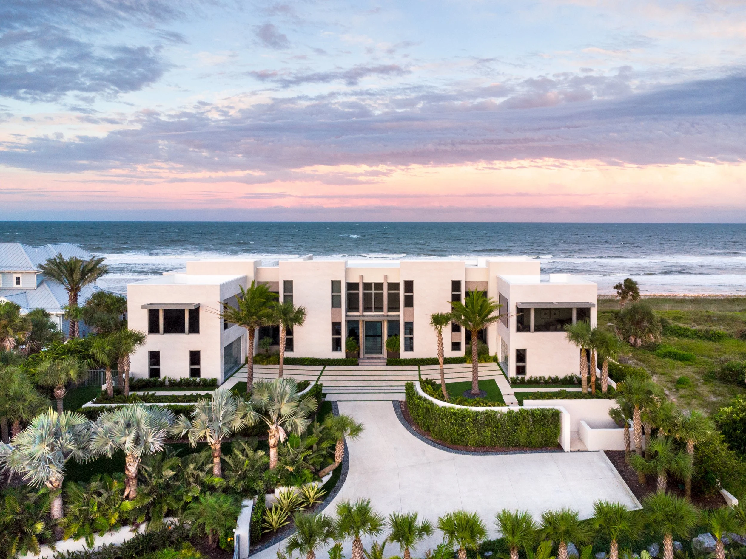 A modern white house with a flat roof, large windows, and palm trees in the front yard, overlooking the ocean with a pink and blue sky overhead.
